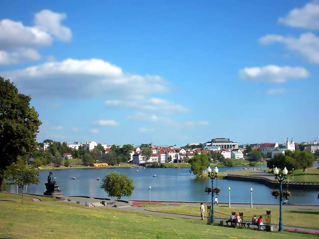 Lake 桌布 壁紙 — 湖邊公園晴朗的一天，人們在長椅上放鬆，船隻在水上，周圍是綠樹和城市建築，藍天白雲。
