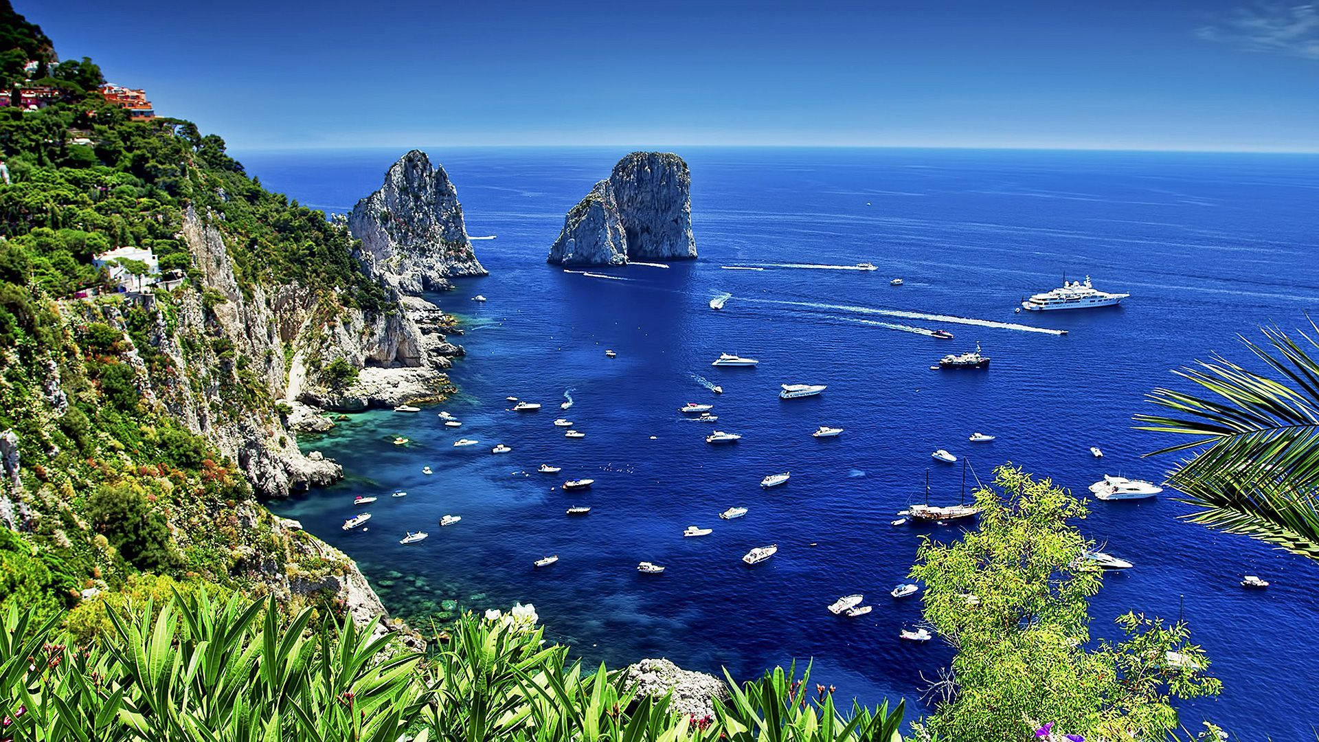 Capri 桌布 壁紙 — 湛藍的海面上點綴著船隻，坐落於鬱鬱蔥蔥的綠色懸崖和標誌性岩層之間，天空晴朗。