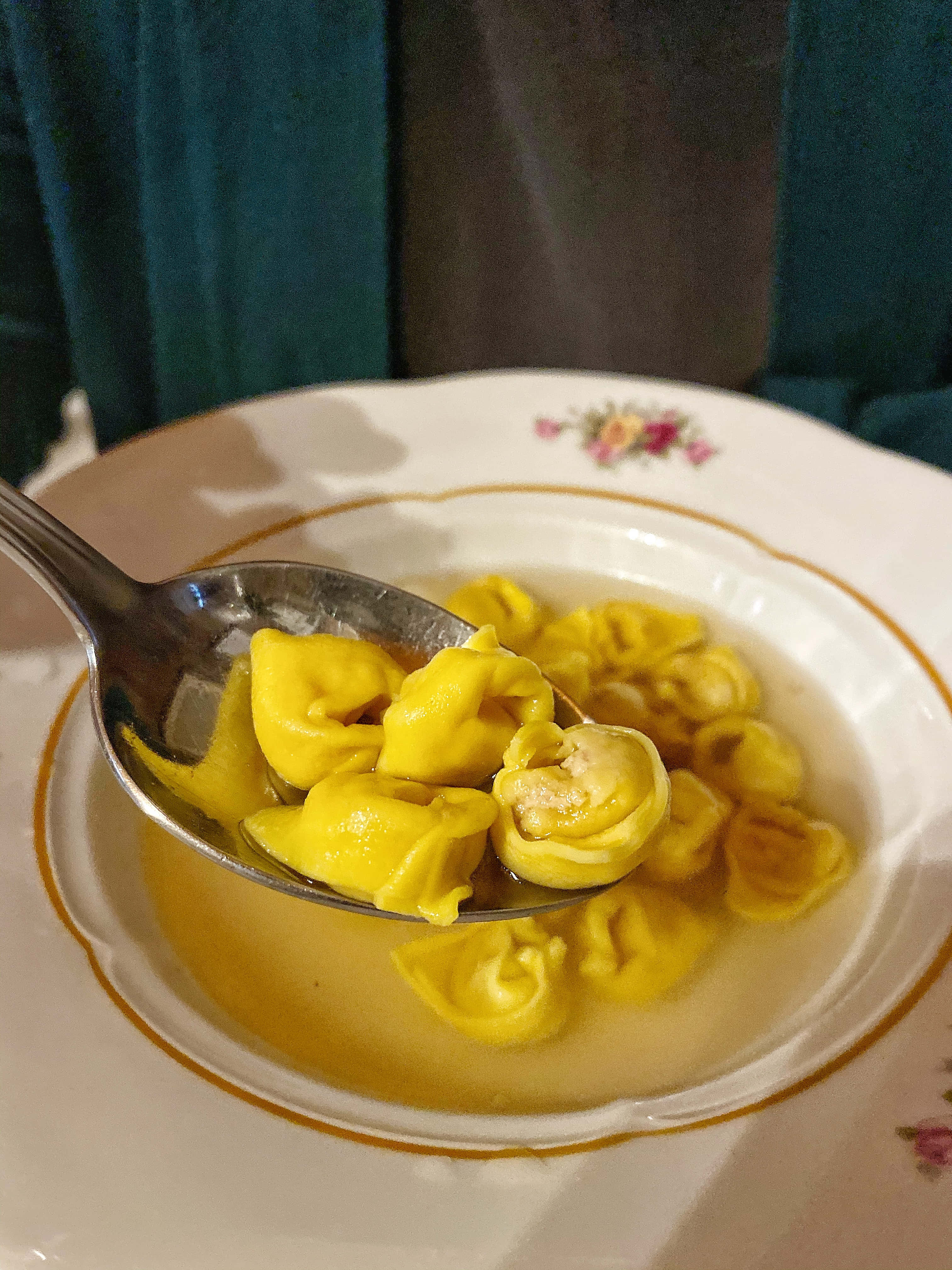 Food Photography 桌布 壁紙 — 湯匙從一個帶有花卉邊緣和金色飾邊的白色碗中，舀起金黃色的義大利餃子（tortellini），浸在奶油湯汁中。