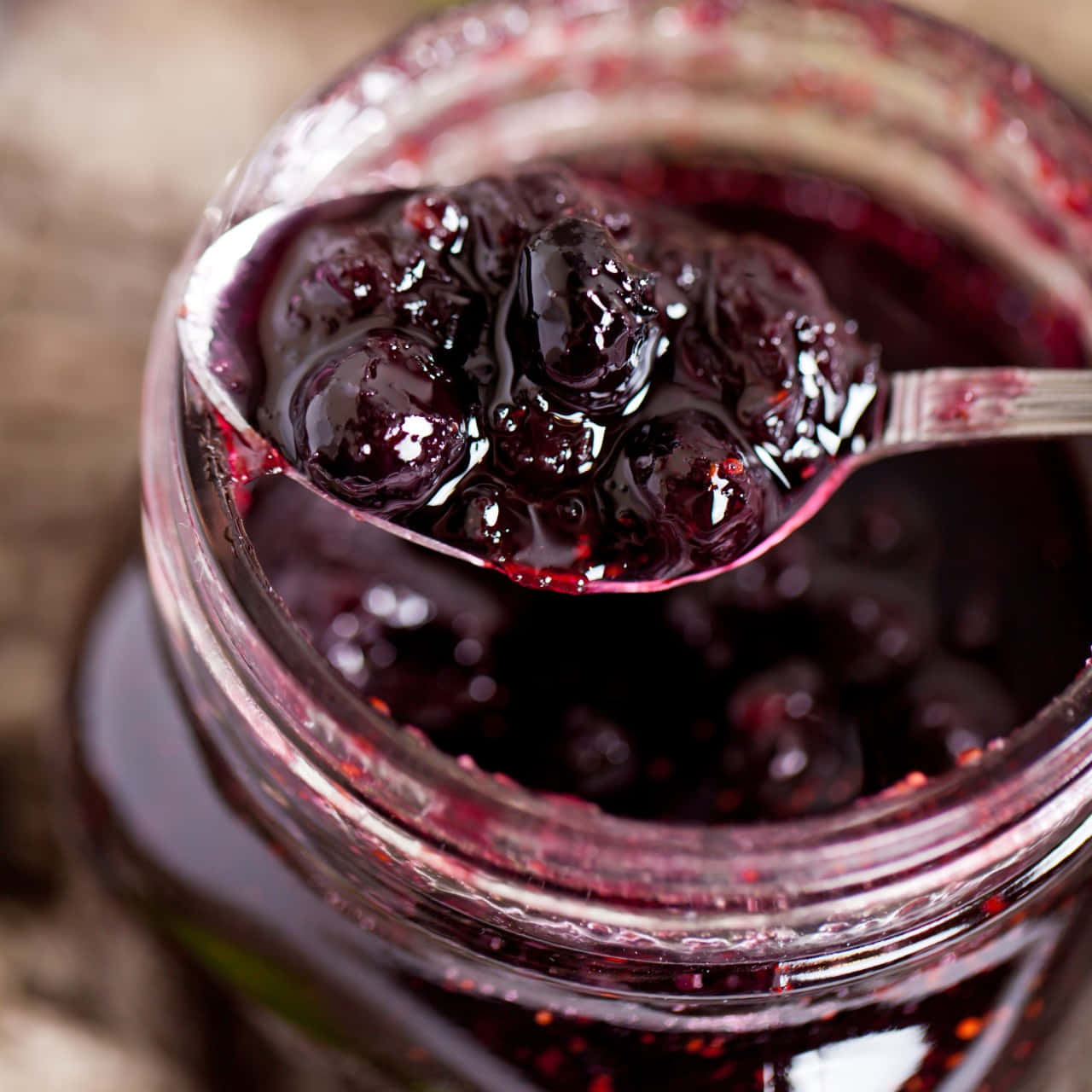 Blueberry Jam 桌布 壁紙 — 湯匙從玻璃罐中舀起閃亮的深紫色藍莓果醬，背景是質樸的木頭。