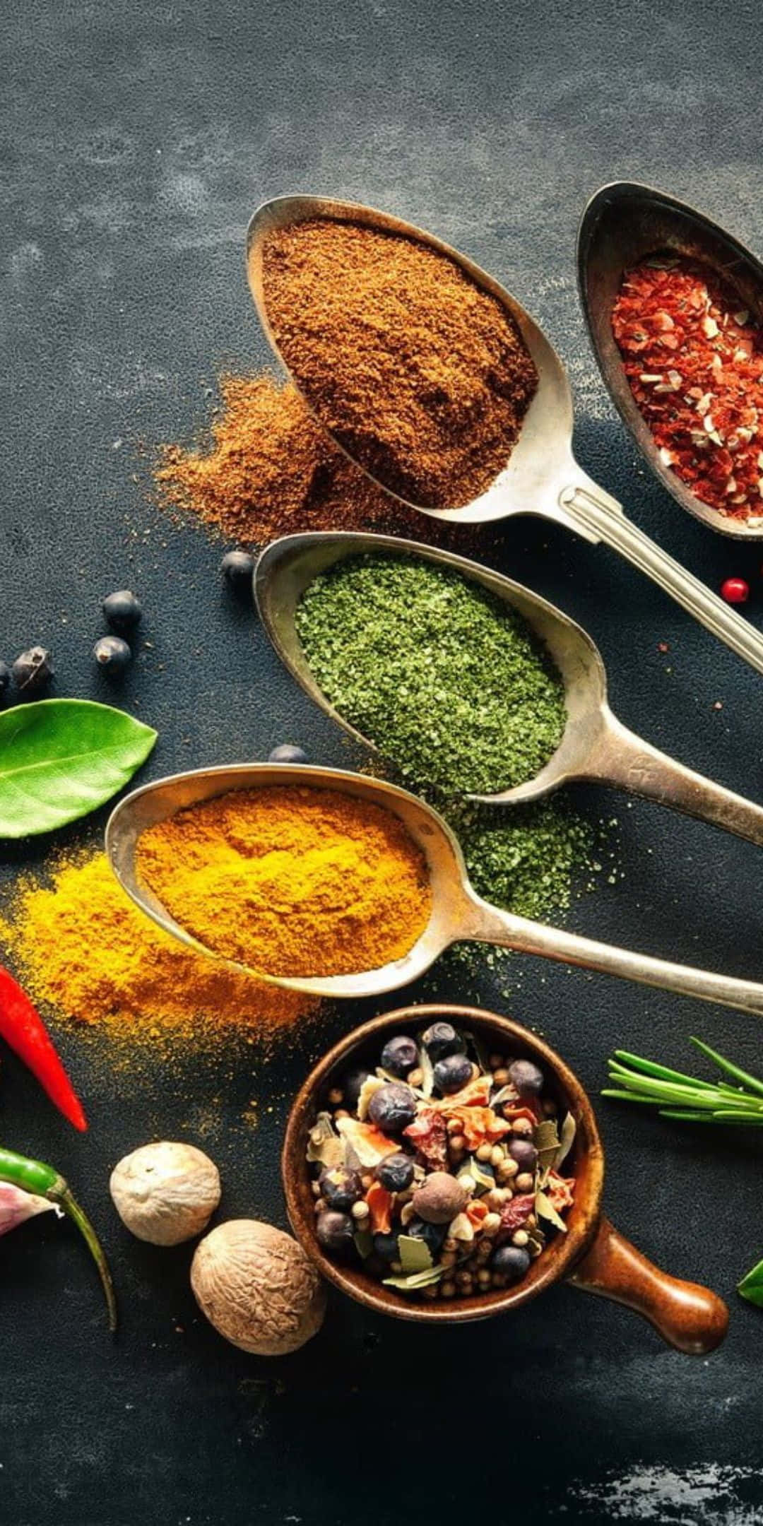 Food Photography 桌布 壁紙 — 湯匙盛滿色彩鮮豔的香料，如薑黃、小茴香和辣椒片，排列在深色紋理表面上，周圍散落著新鮮香草和整粒香料。
