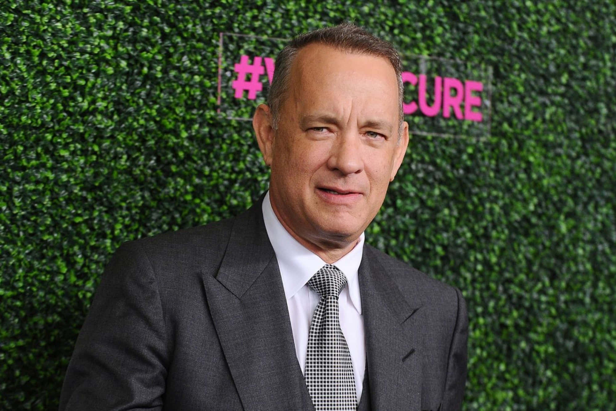 Celebrity 桌布 壁紙 — 湯姆漢克斯身穿灰色西裝和格紋領帶，站在綠色樹籬牆前，牆上有粉紅色的hashtag cure標誌。