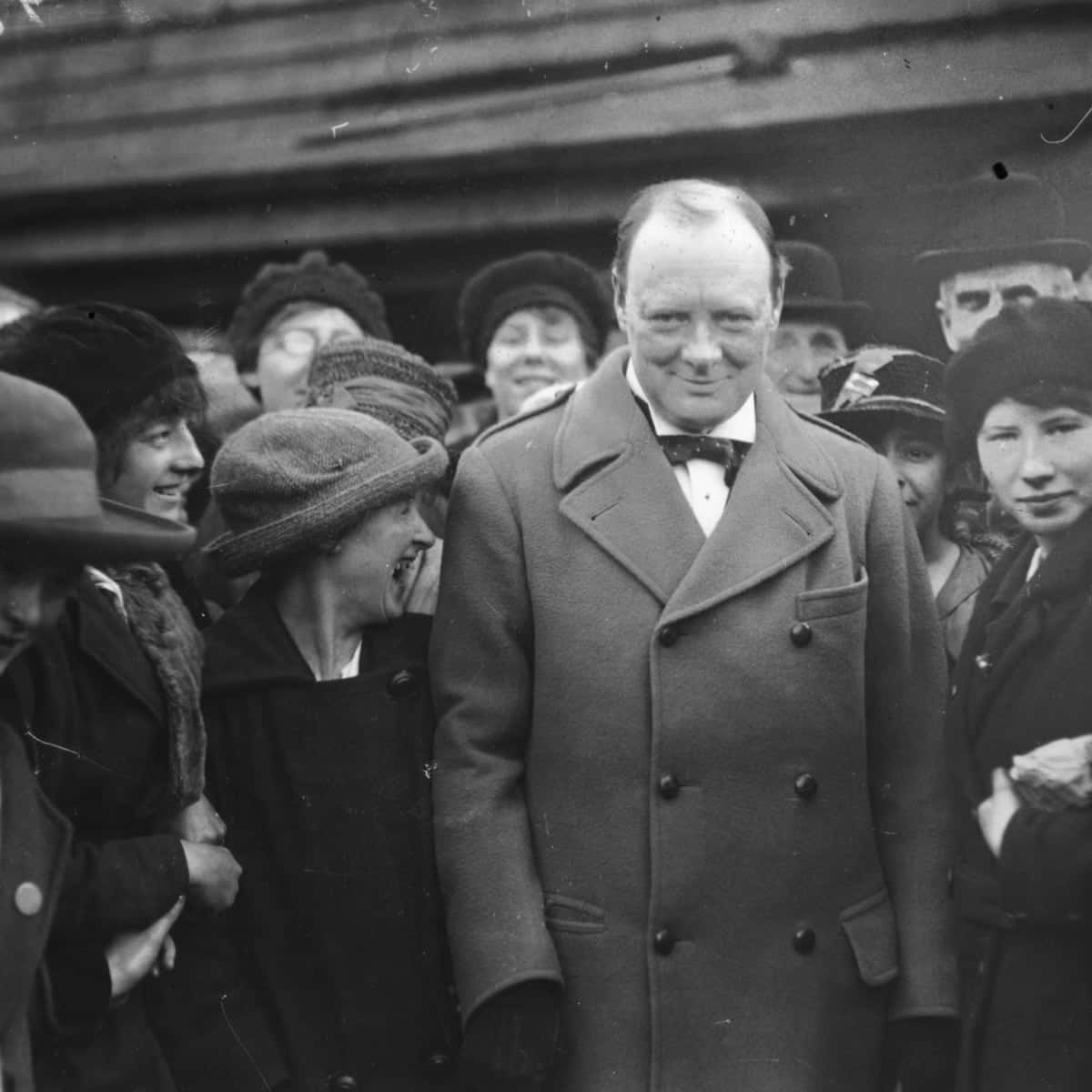 Winston Churchill 桌布 壁紙 — 溫斯頓·邱吉爾面帶溫暖笑容，站在一群穿著復古冬季大衣和帽子的微笑旁觀者面前。