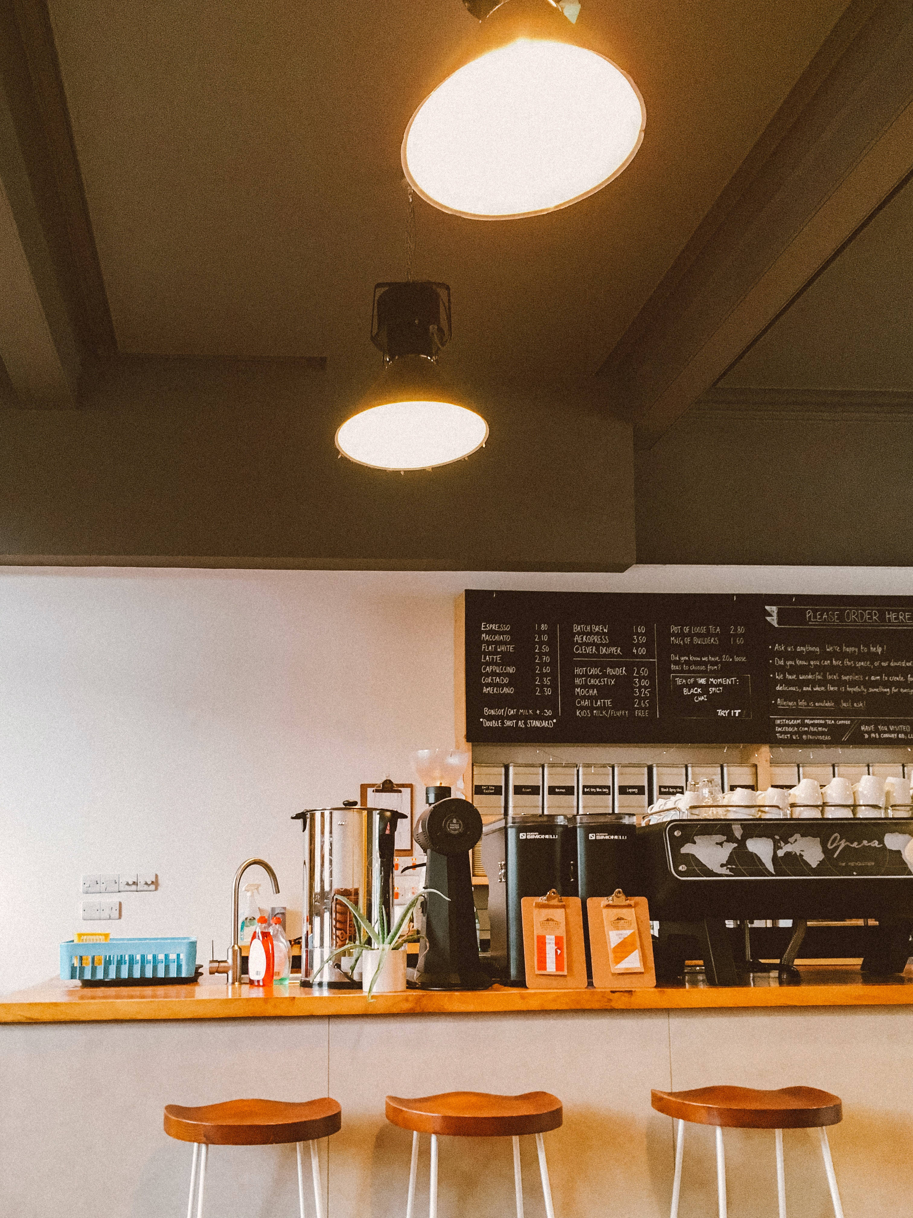 Coffee Shop 桌布 壁紙 — 溫暖照明的咖啡店內部，設有木製吧檯凳和黑板菜單，下方是裸露的天花板橫樑。