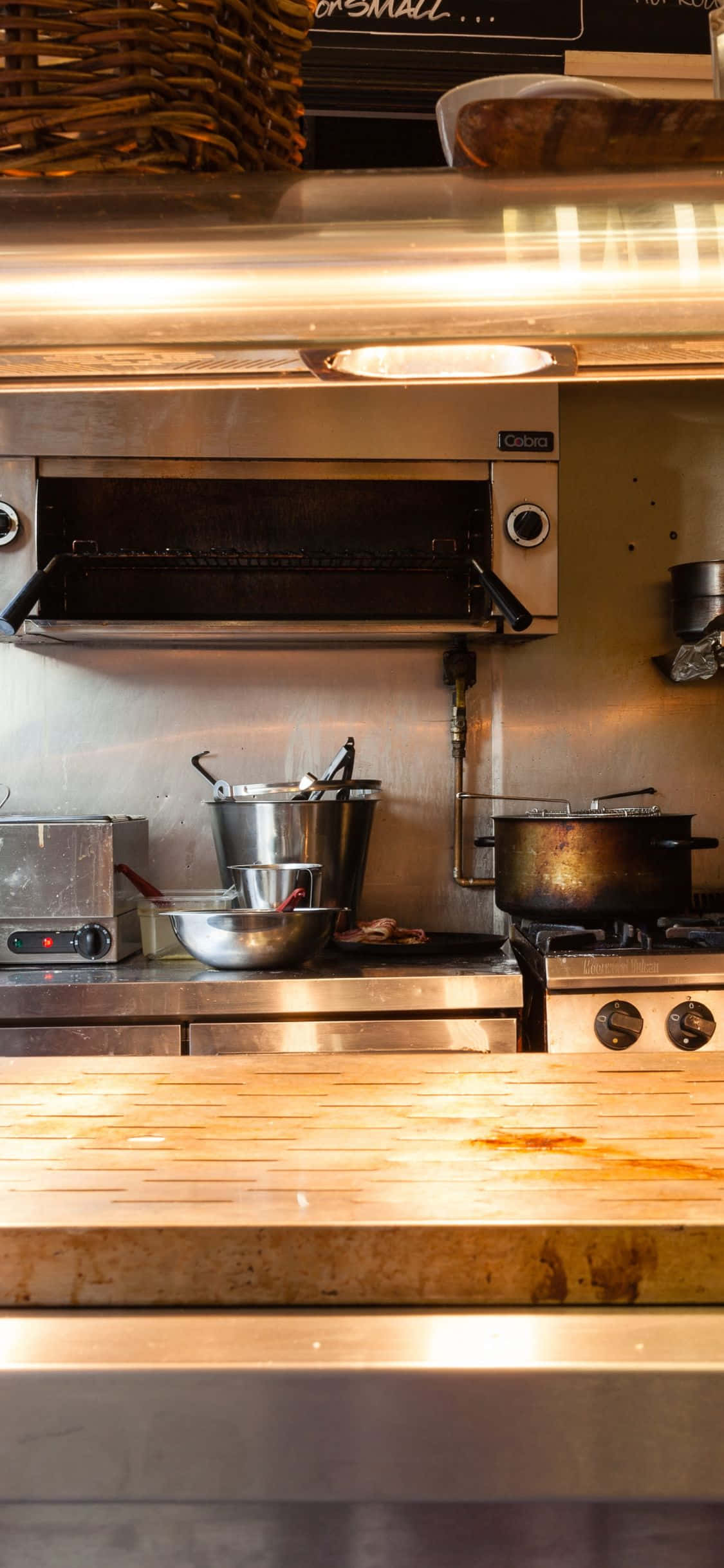 kitchen 桌布 壁紙 — 溫暖照明的商業廚房，配有不鏽鋼器具和木製檯面，顯示使用痕跡。