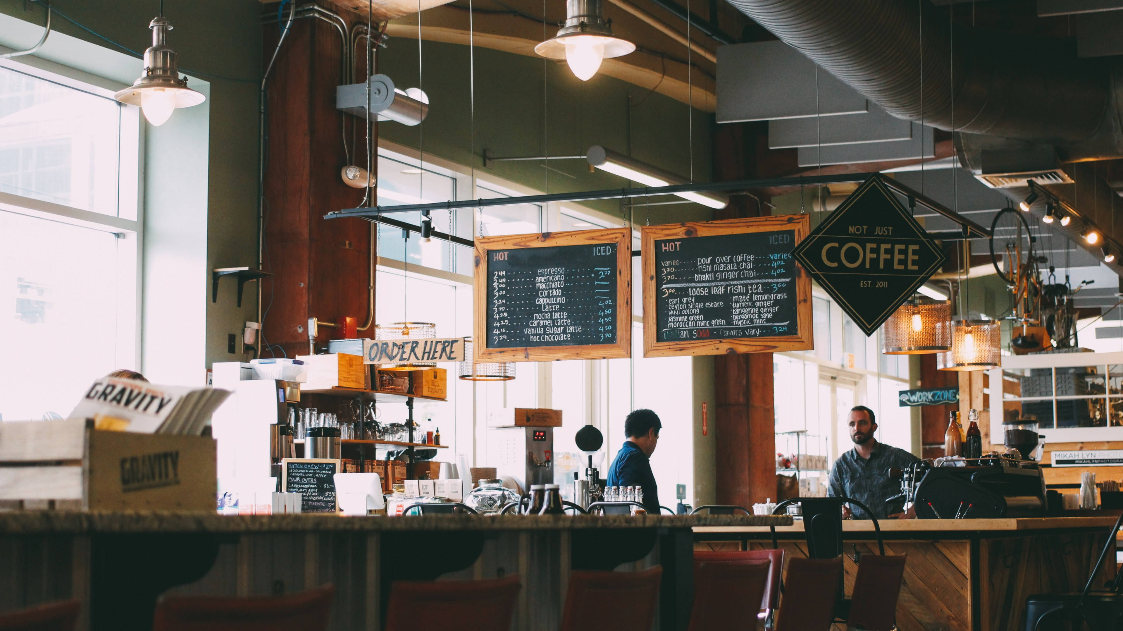 Coffee Shop 桌布 壁紙 — 溫暖照明的工業風咖啡店，有外露管道、木製櫃檯、黑板菜單，以及櫃檯後工作的咖啡師。