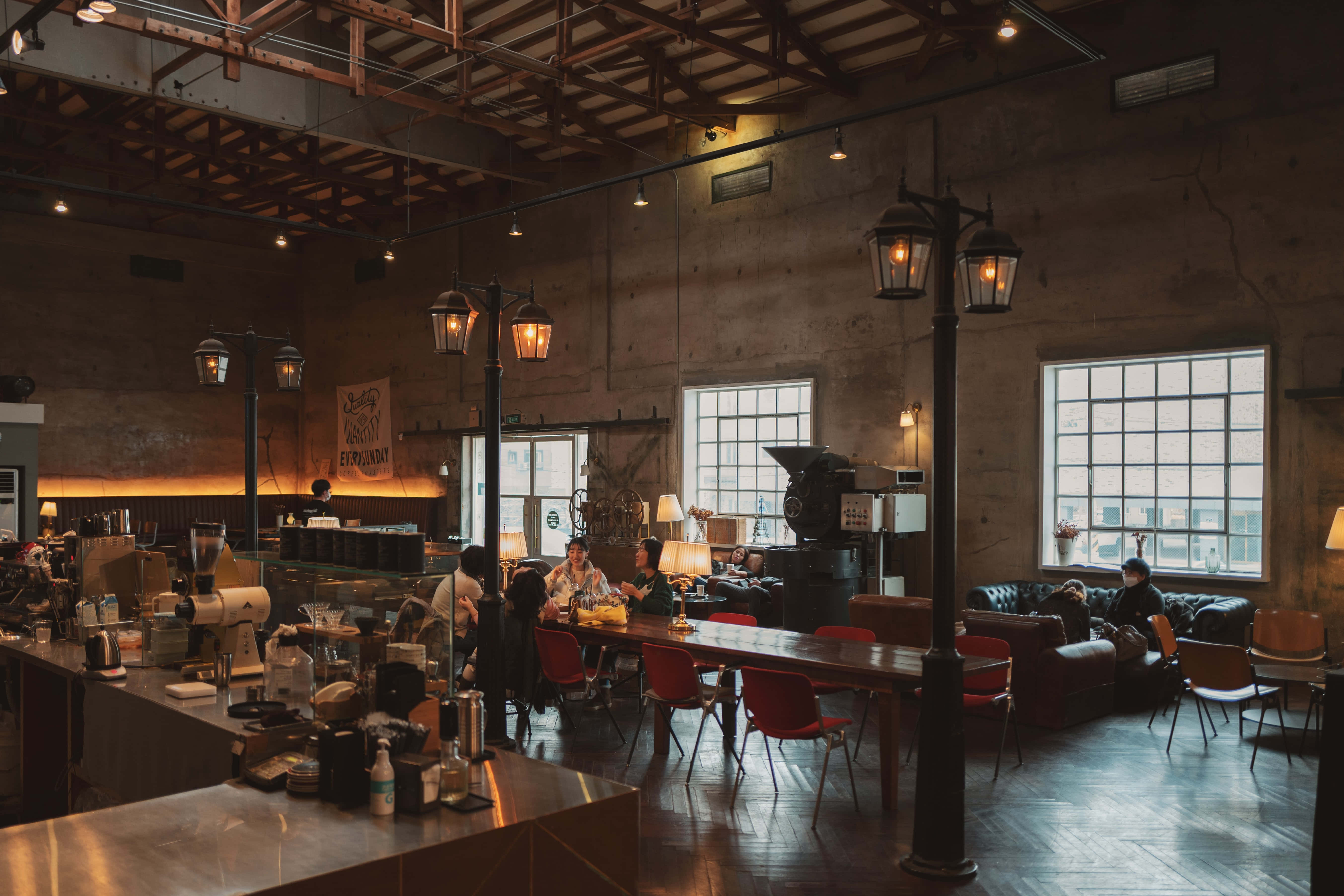 Coffee Aesthetic 桌布 壁紙 — 溫暖照明的工業風咖啡店，有裸露橫樑、顧客在餐桌旁，以及復古燈籠下的紅色椅子。