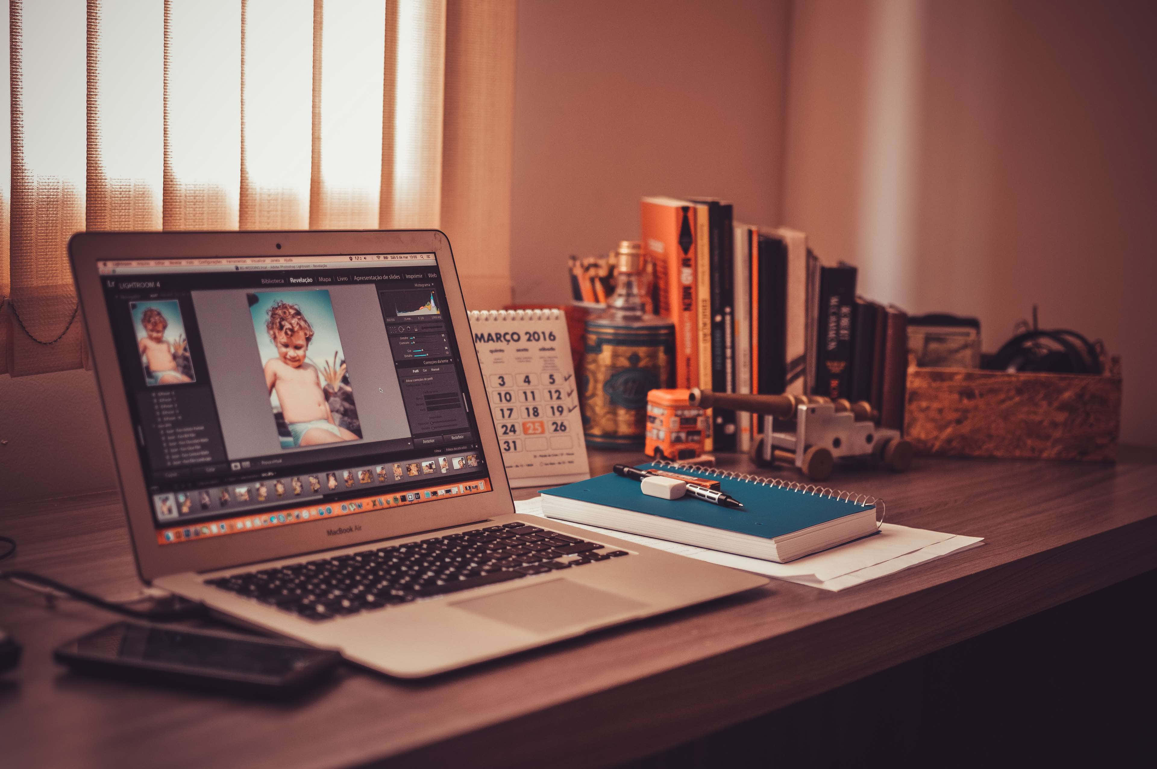 Working From Home 桌布 壁紙 — 溫暖照明的木製書桌，上面有顯示照片編輯軟體的筆記型電腦，旁邊有日曆筆記本和書籍。