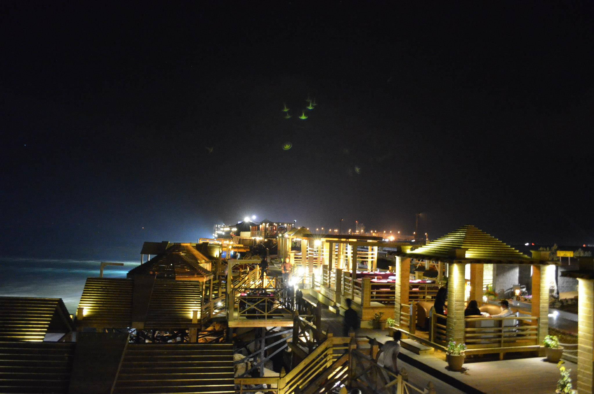 Karachi 桌布 壁紙 — 溫暖照明的木製海濱建築在漆黑的夜空中閃耀，遠方城市燈光與下方海浪相伴。