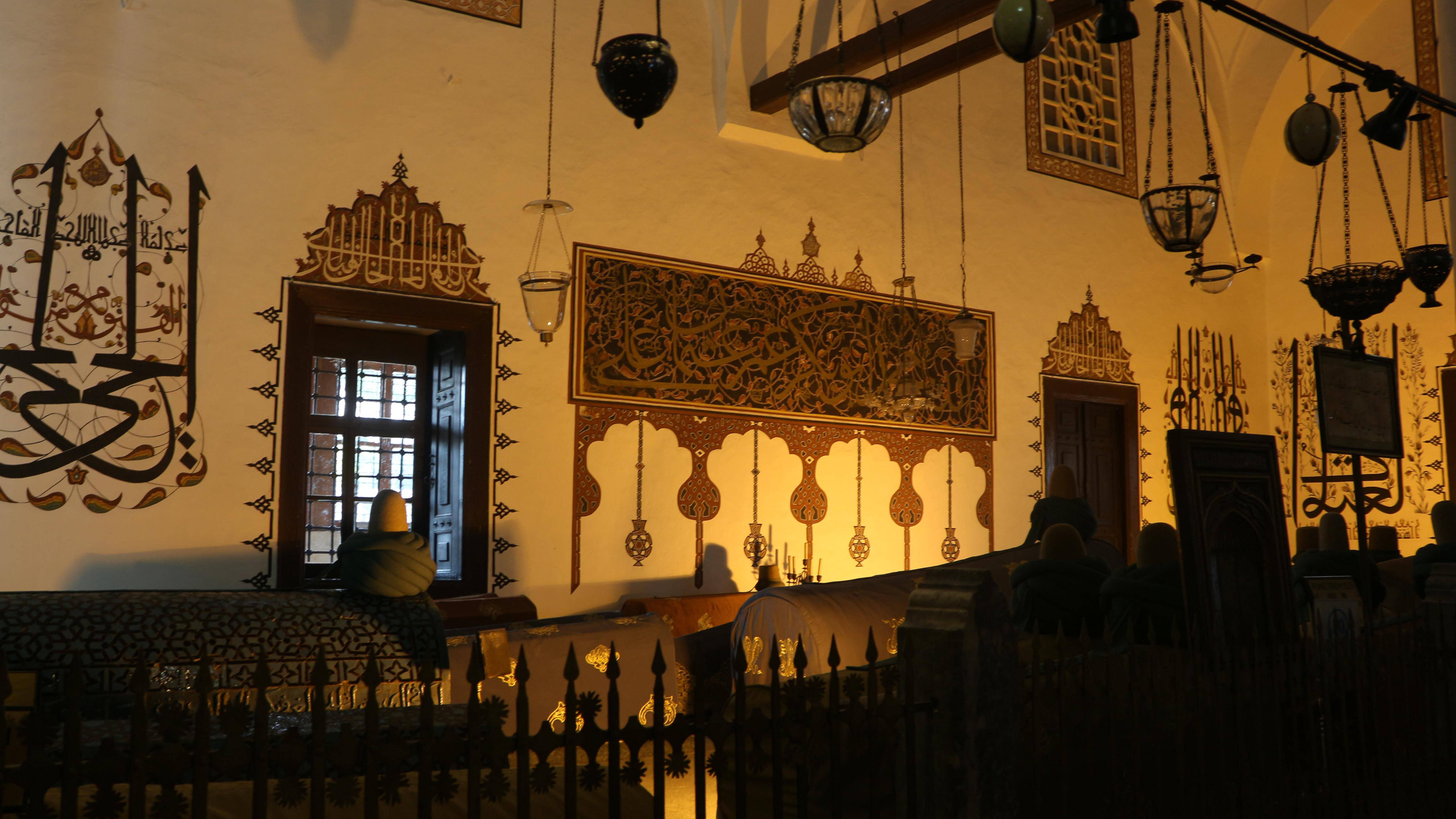 Konya 桌布 壁紙 — 溫暖照明的清真寺內部，帶有複雜的阿拉伯式牆壁雕刻、懸掛燈籠和剪影中的禮拜者。