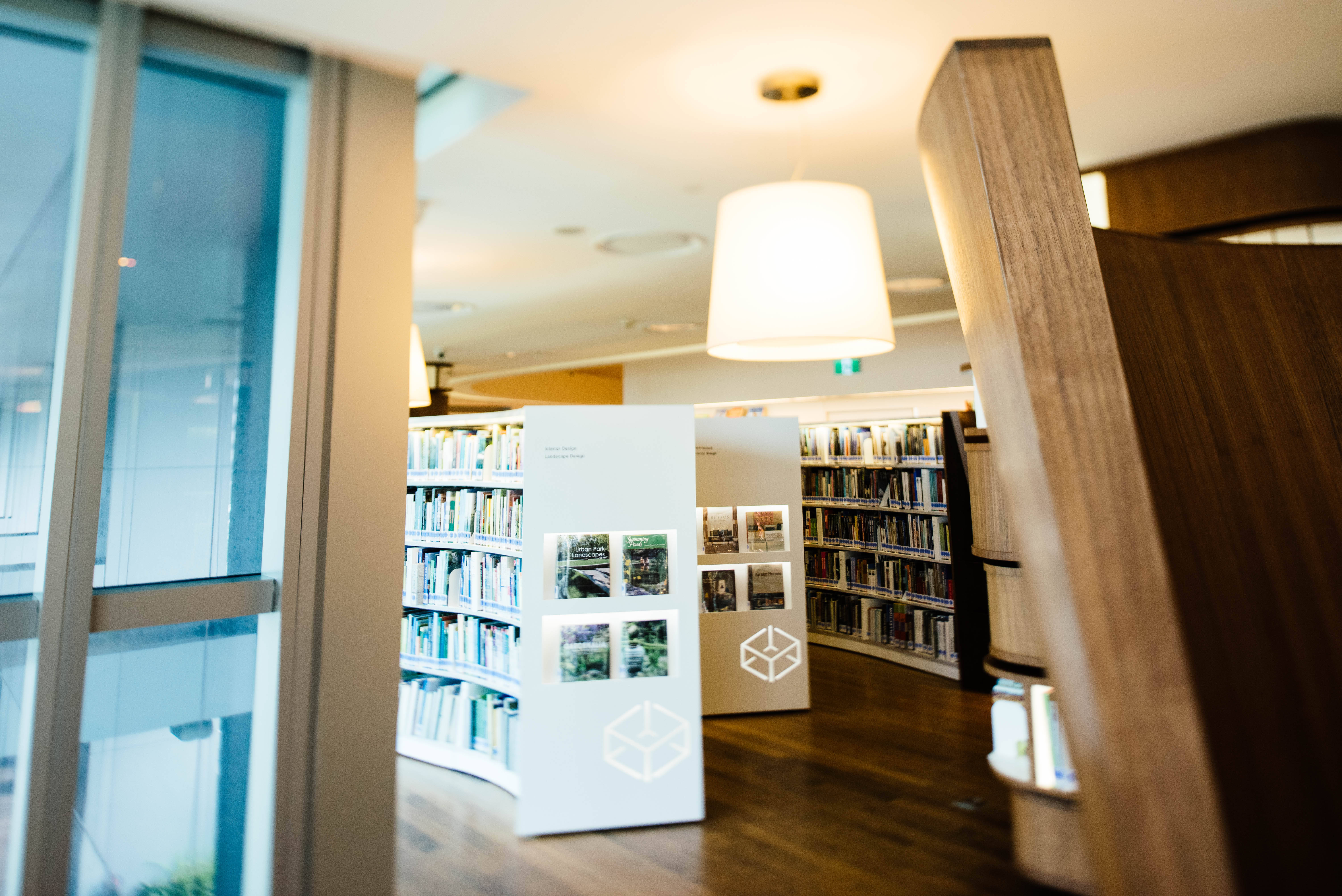 Shelves 桌布 壁紙 — 溫暖照明的現代圖書館內部，木地板、書架和一個展示照片展品的白色展示架。