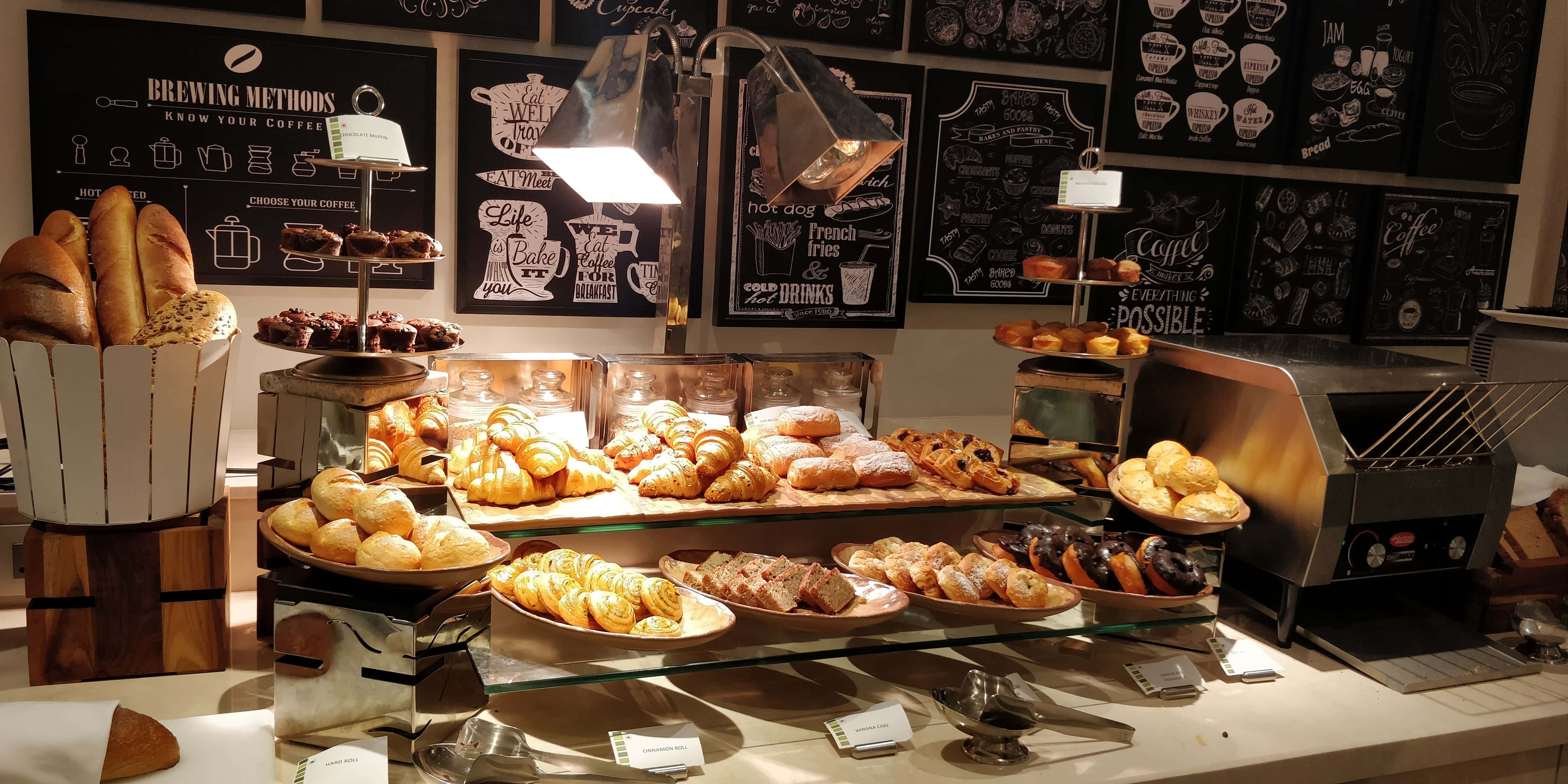 Bakery 桌布 壁紙 — 溫暖照明的麵包店展示櫃，在黑板上菜單下堆滿了金黃色的牛角包、丹麥酥和新鮮麵包。