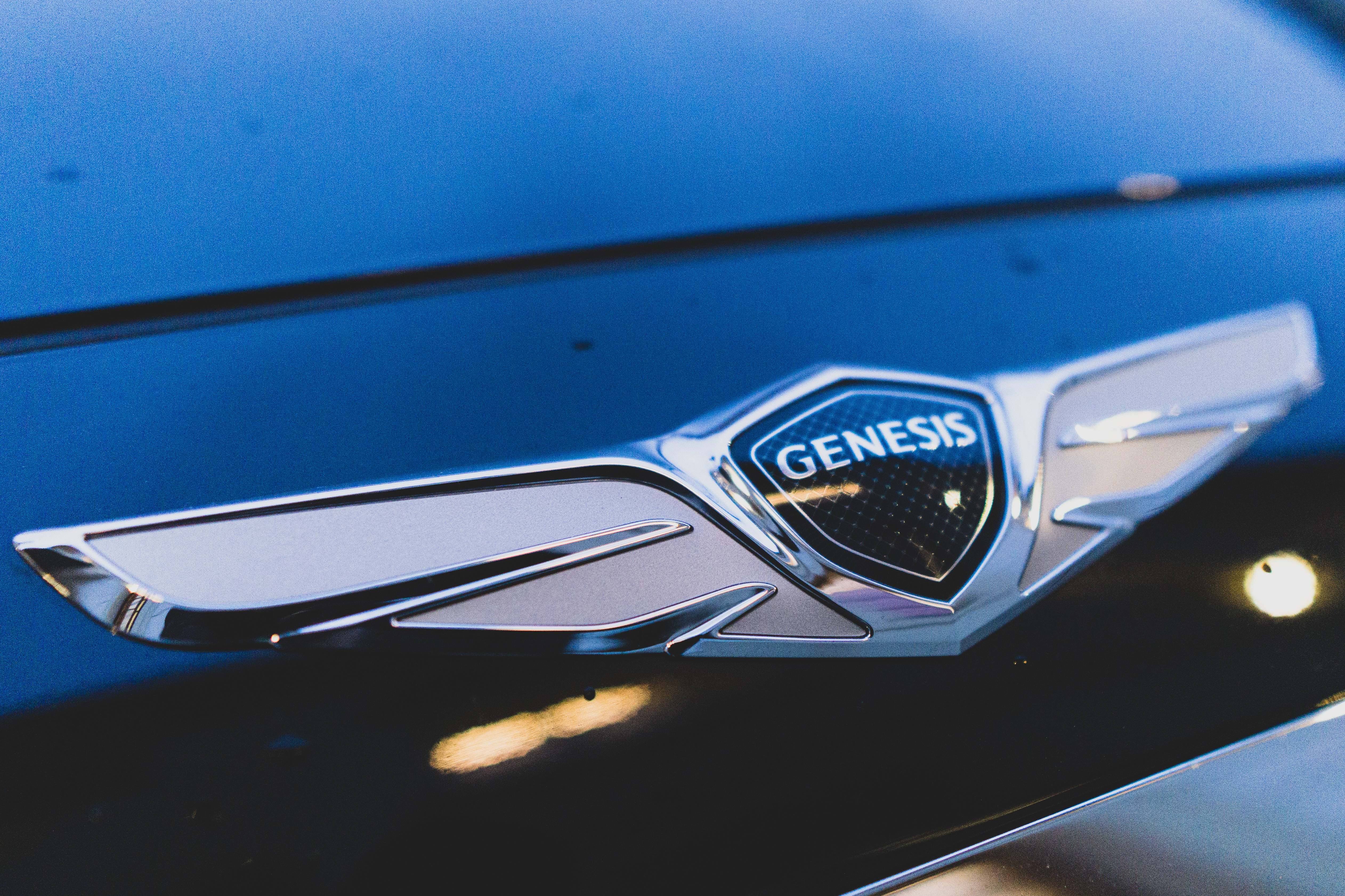 Genesis 桌布 壁紙 — 溫暖燈光下，光滑藍色引擎蓋上閃亮鍍鉻Genesis汽車標誌的特寫。