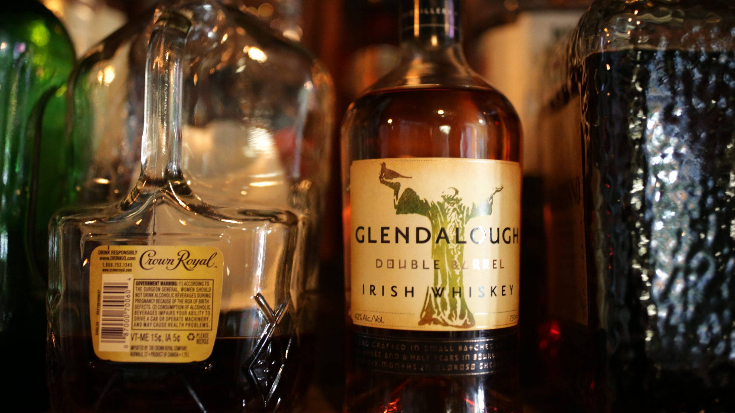 節日 桌布 壁紙 — 溫暖燈光下威士忌酒瓶的特寫，包括Glendalough和Crown Royal，放在酒吧架上，玻璃器皿模糊。