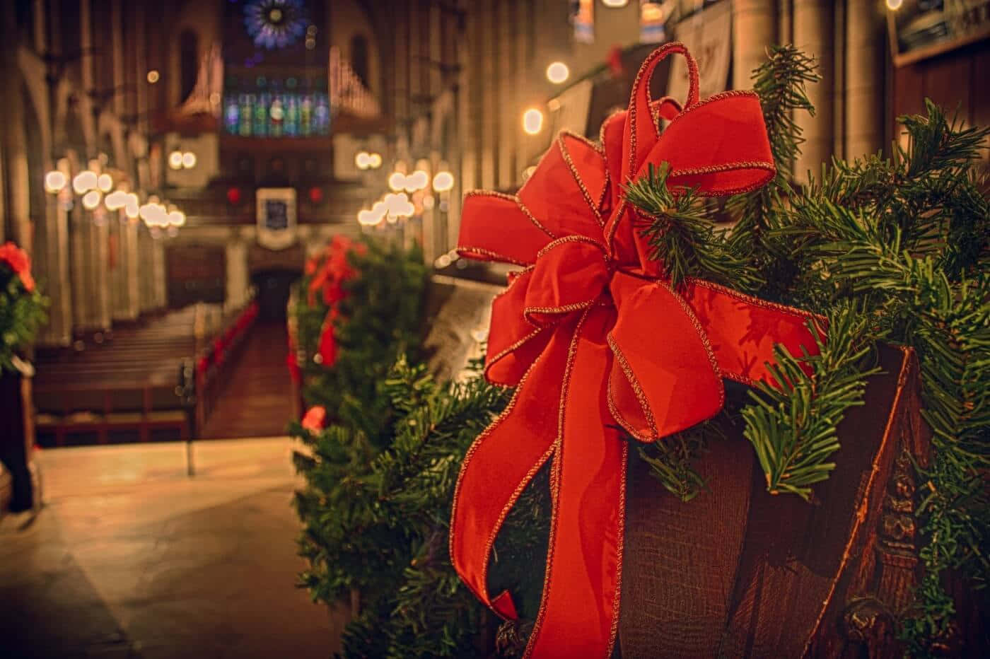 Religious Christmas 桌布 壁紙 — 溫暖燈光下的大教堂走廊中，綠色花環上的一個節日紅色蝴蝶結特寫。