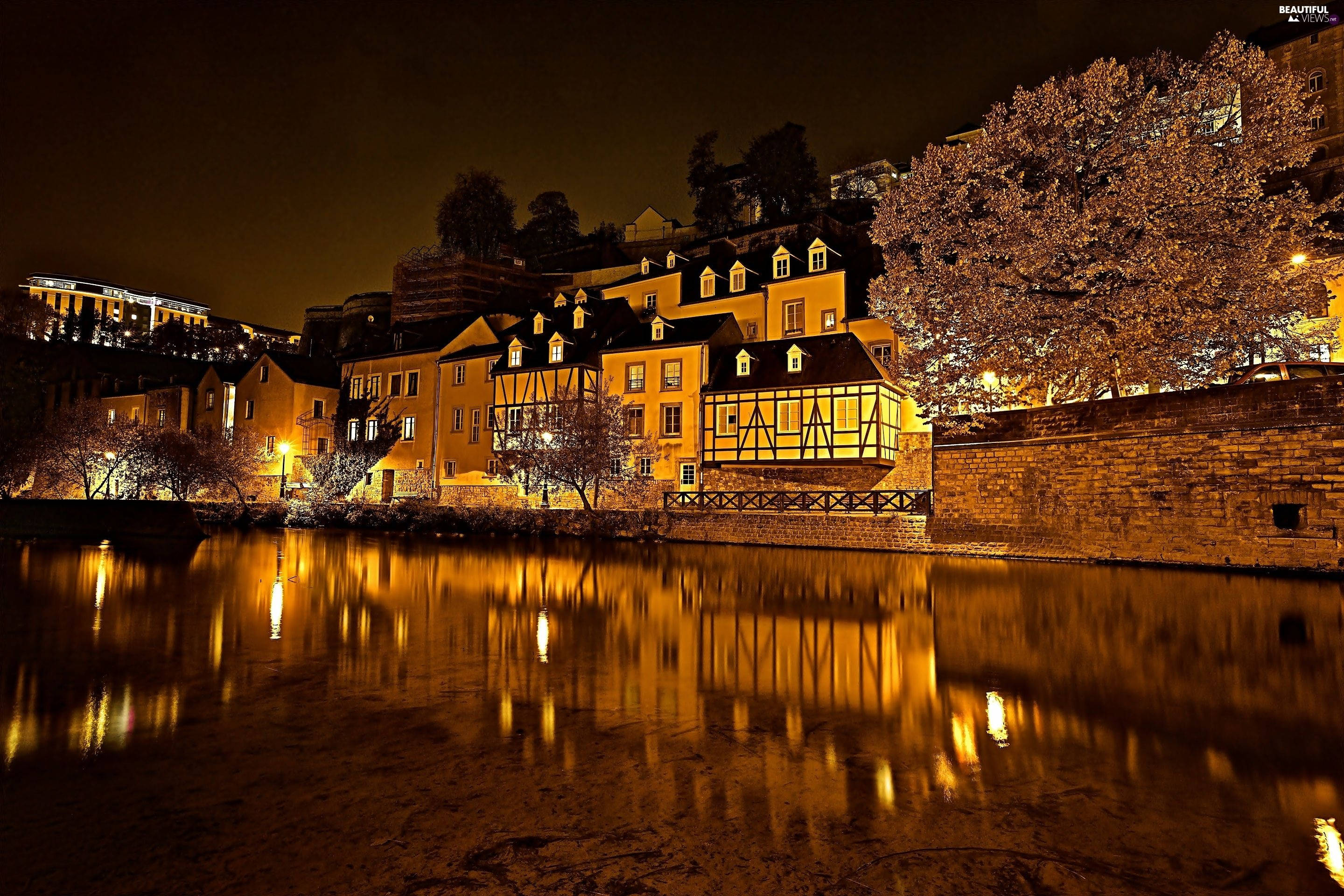 Luxembourg 桌布 壁紙 — 溫暖燈光照亮半木結構房屋，在黑暗夜空下倒映在平靜水面上，附近樹木閃爍光芒。