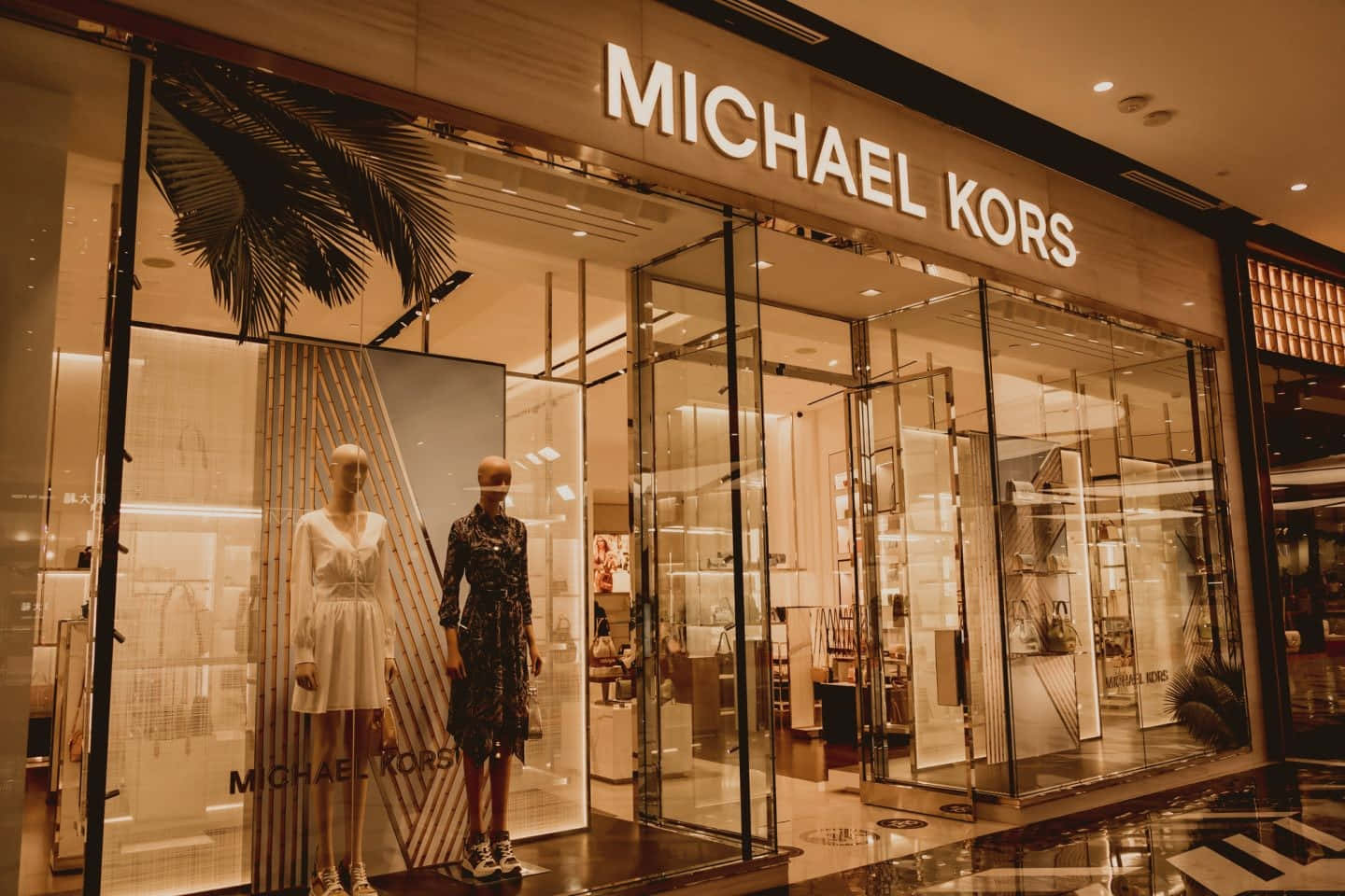 麥可·寇斯 桌布 壁紙 — 溫暖燈光的Michael Kors店鋪正面，時尚連衣裙的人體模型和棕櫚葉裝飾在拋光地板上反射。