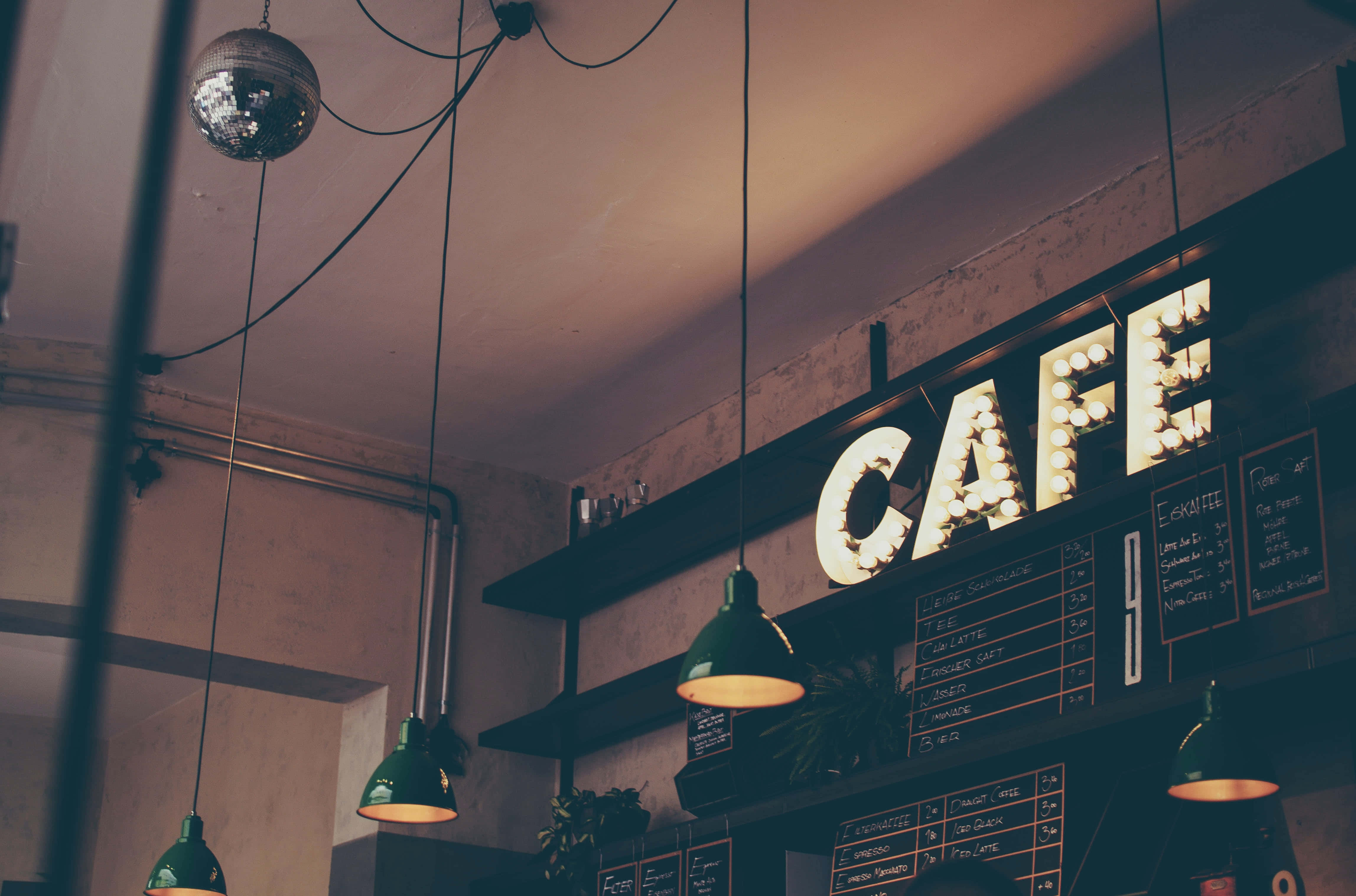Coffee Aesthetic 桌布 壁紙 — 溫暖燈光的咖啡館內部，發光的招牌懸掛在黑板上方，搭配復古綠色吊燈。