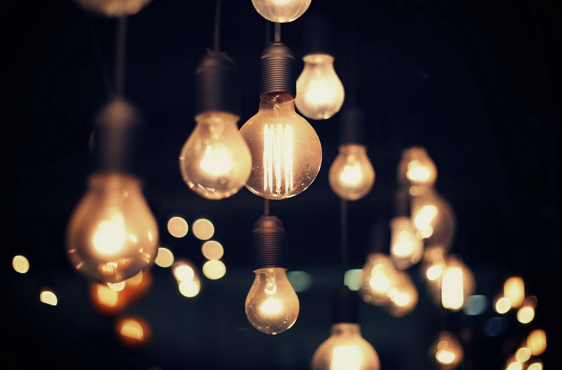 Light Bulb 桌布 壁紙 — 溫暖發光的燈絲燈泡懸掛在黑暗背景中，遠處有柔和的散景燈光。