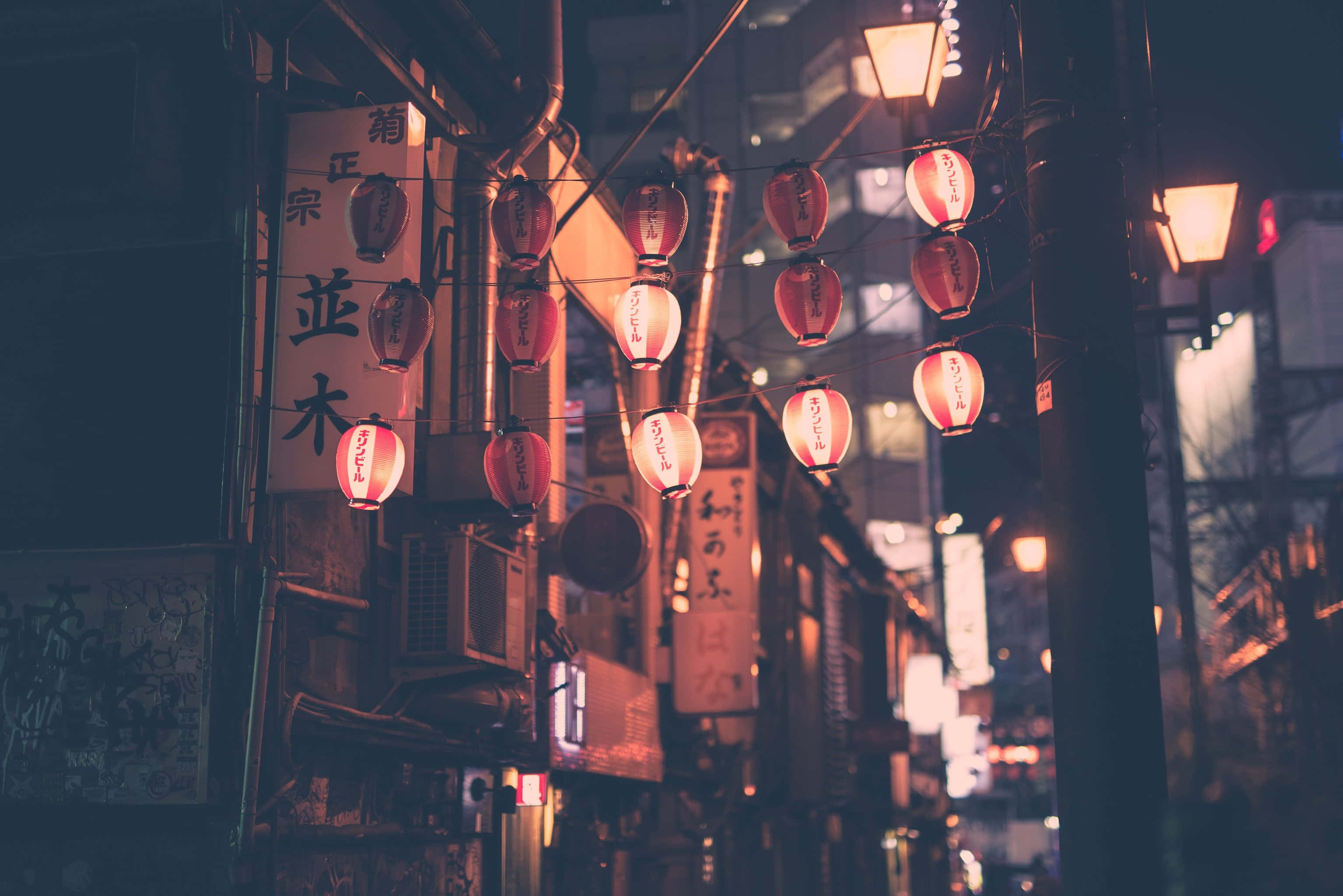 Red Japanese 桌布 壁紙 — 溫暖發光的紅色燈籠掛在狹窄的日本小巷中，夜晚時分，柔和的光線灑在風化的建築物和招牌上。