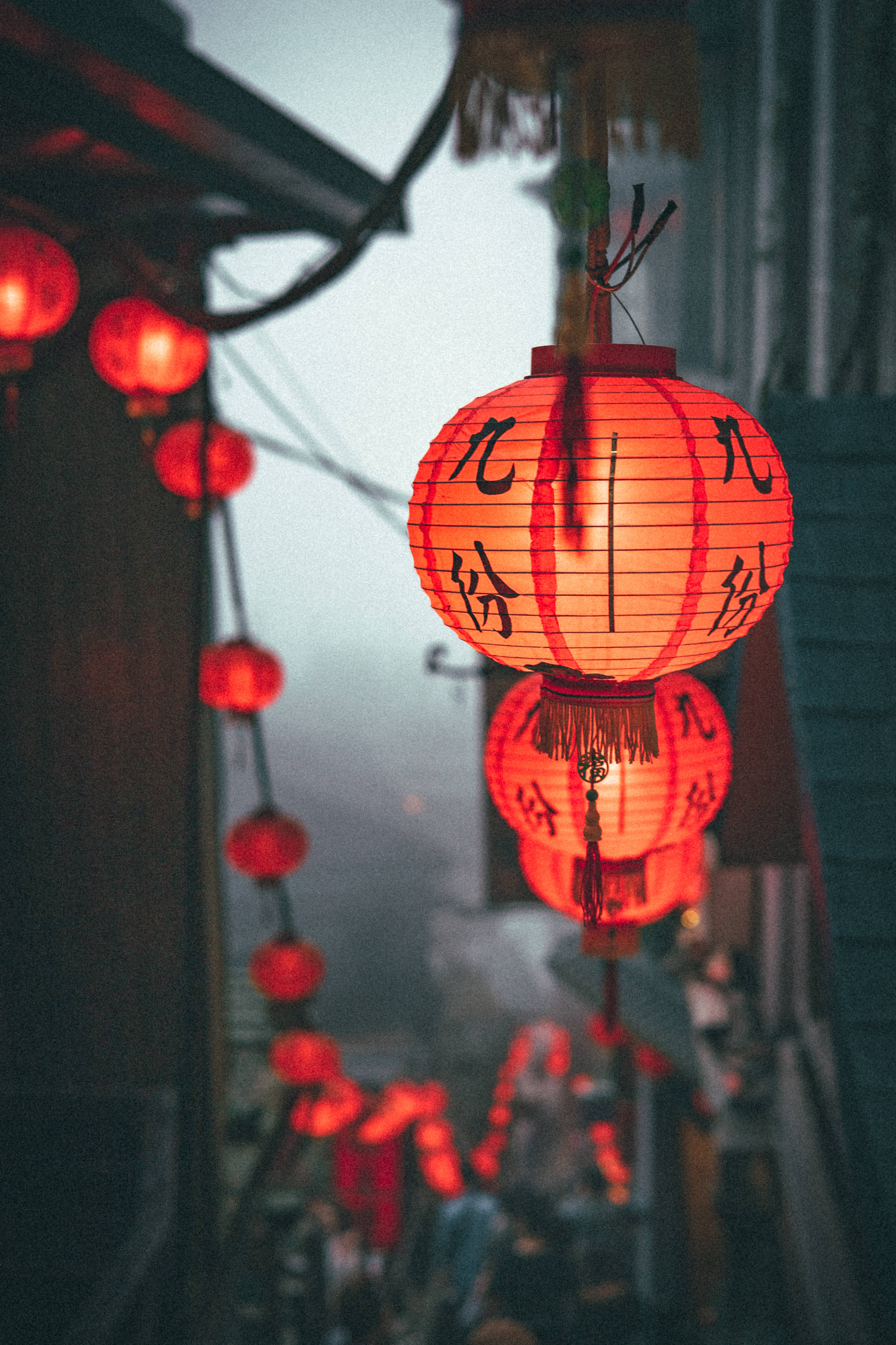 中文 桌布 壁紙 — 溫暖發光的紅色紙燈籠，帶有黑色書法，懸掛在黃昏時分霧氣瀰漫的小巷中。