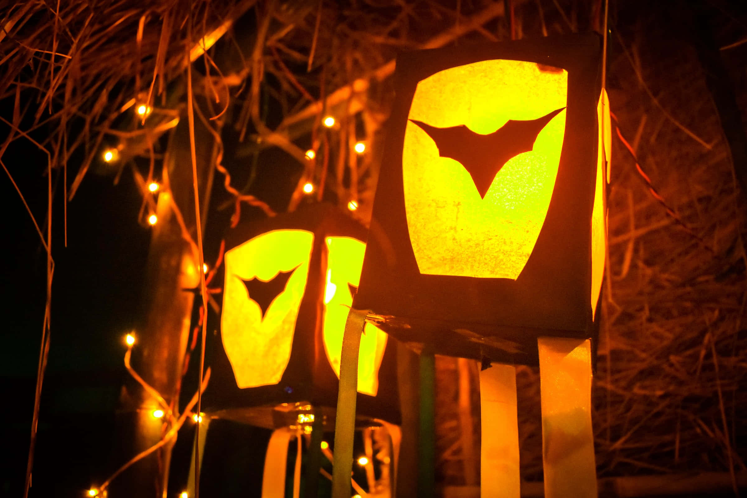Halloween Desktop 桌布 壁紙 — 溫暖發光的蝙蝠形燈籠掛在樹枝和仙女燈之間，處於黑暗的夜晚環境中。