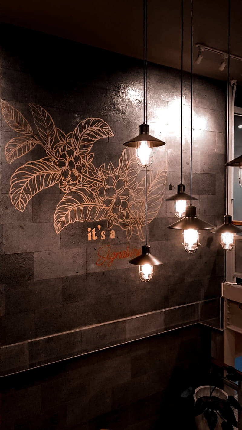 Coffee Shop 桌布 壁紙 — 溫暖的吊燈在舒適的咖啡館中，對著裝飾有發光咖啡植物壁畫的深色石牆閃耀。