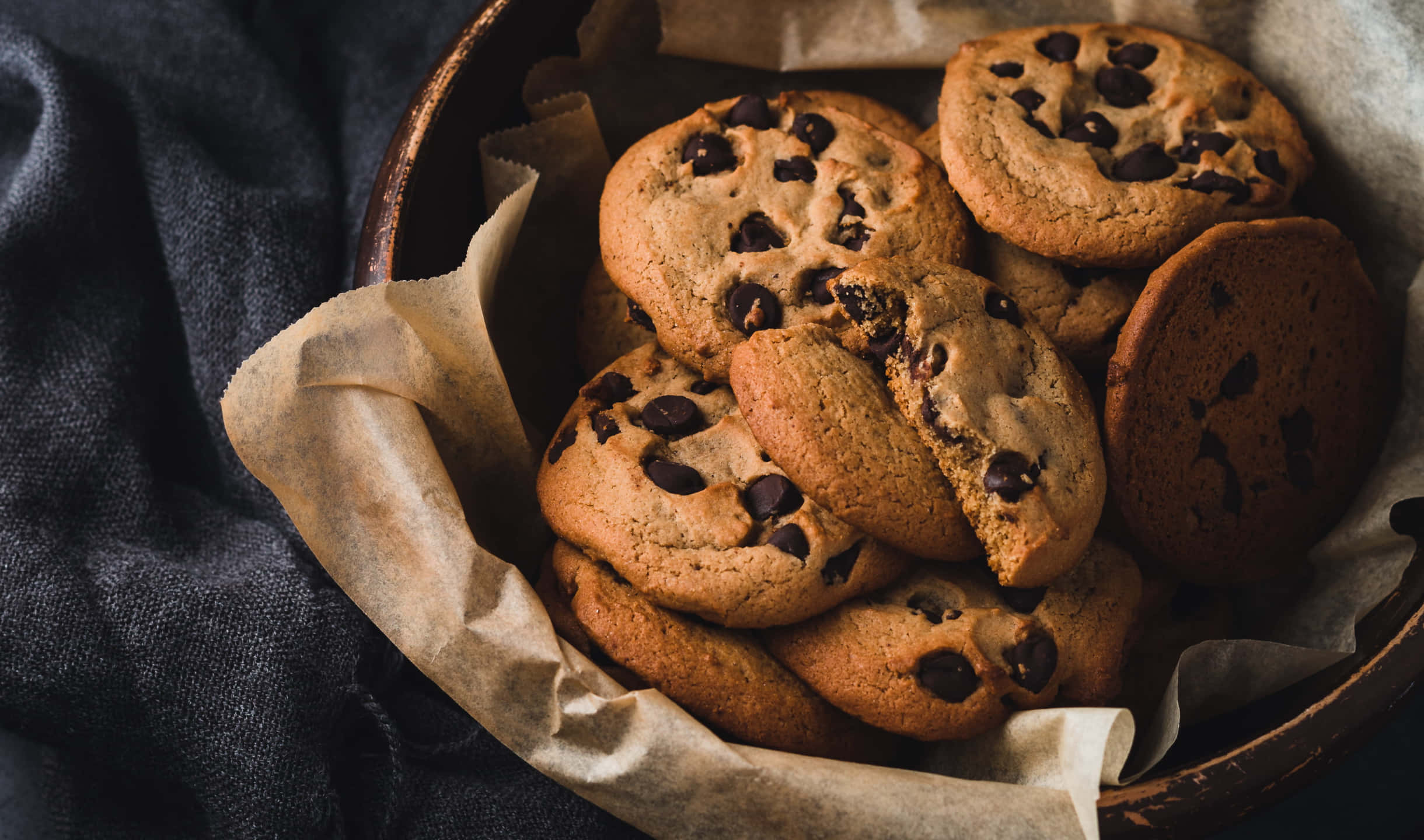 Cookies 桌布 壁紙 — 溫暖的巧克力餅乾堆疊在鋪有烘焙紙的鄉村風格木碗中，置於深色紋理布上。