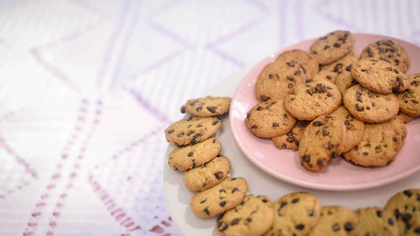 1366x768 Cookies 桌布 壁紙 — 溫暖的巧克力餅乾從粉紅色盤子溢出到白色圖案桌布上，在柔和日光中。