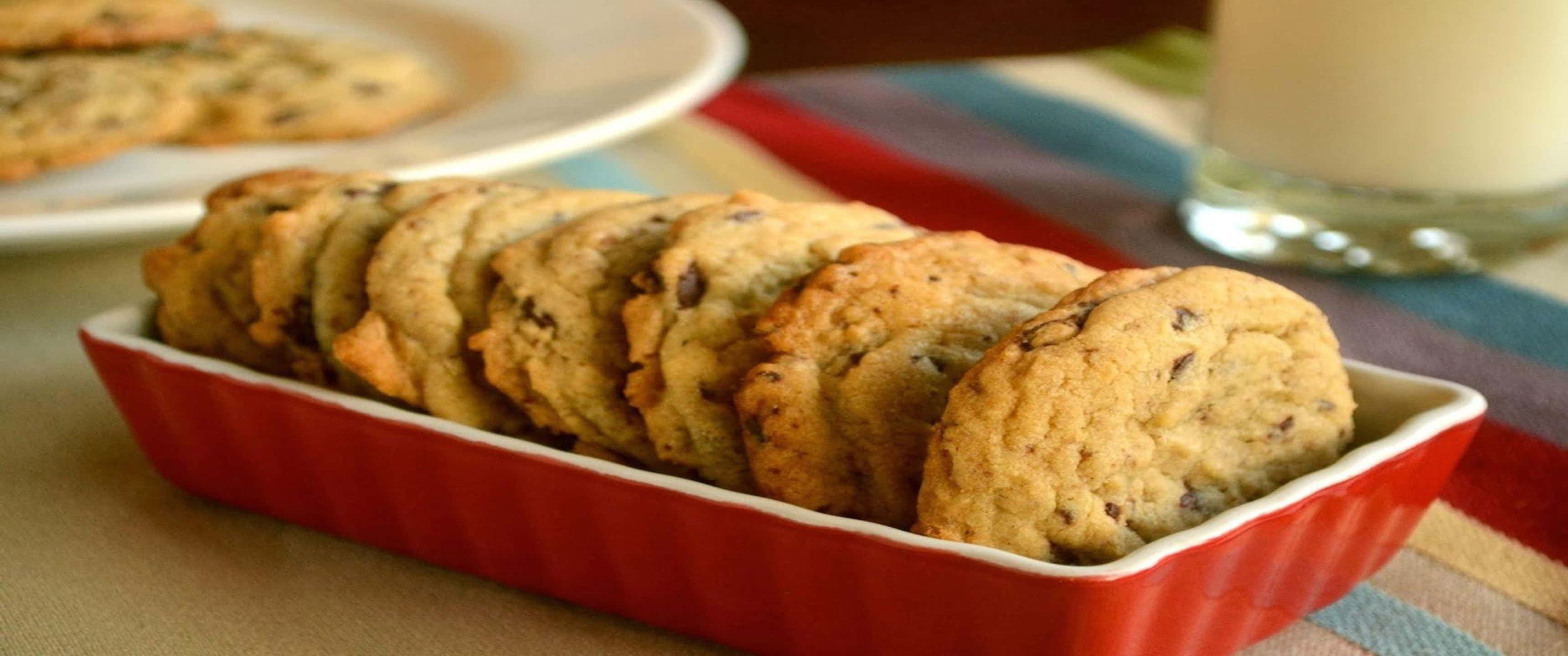 Biscuit 桌布 壁紙 — 溫暖的巧克力餅乾放在紅色陶瓷盤中，旁邊有一杯牛奶，置於條紋布上。