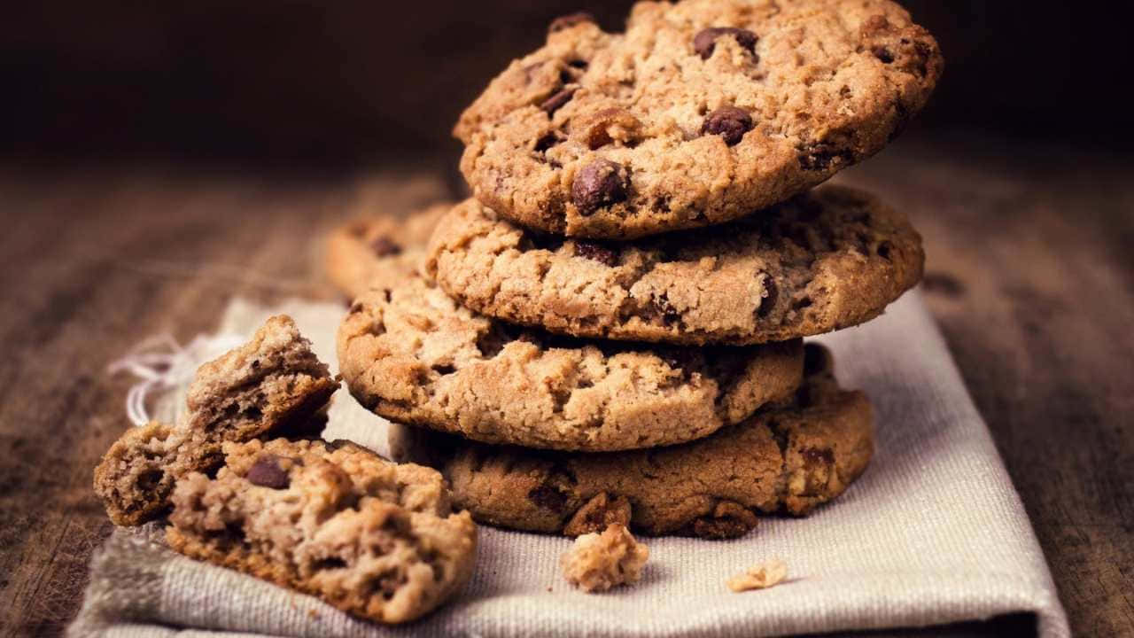 720p Cookies 桌布 壁紙 — 溫暖的巧克力餅乾誘人地堆疊在亞麻餐巾上，麵包屑散落在質樸的木質表面上。