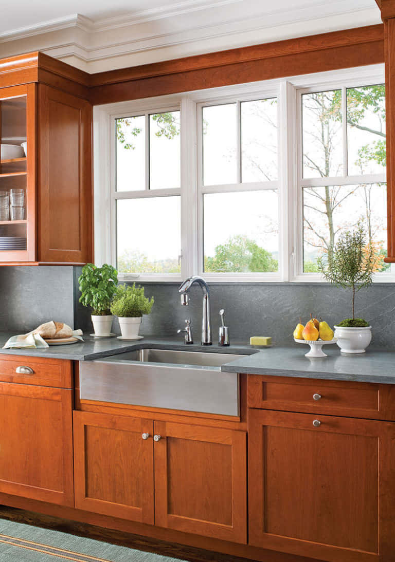 房屋 桌布 壁紙 — 溫暖的木製廚房櫥櫃環繞著不鏽鋼水槽，上方是明亮的窗戶，窗外有綠色植物。