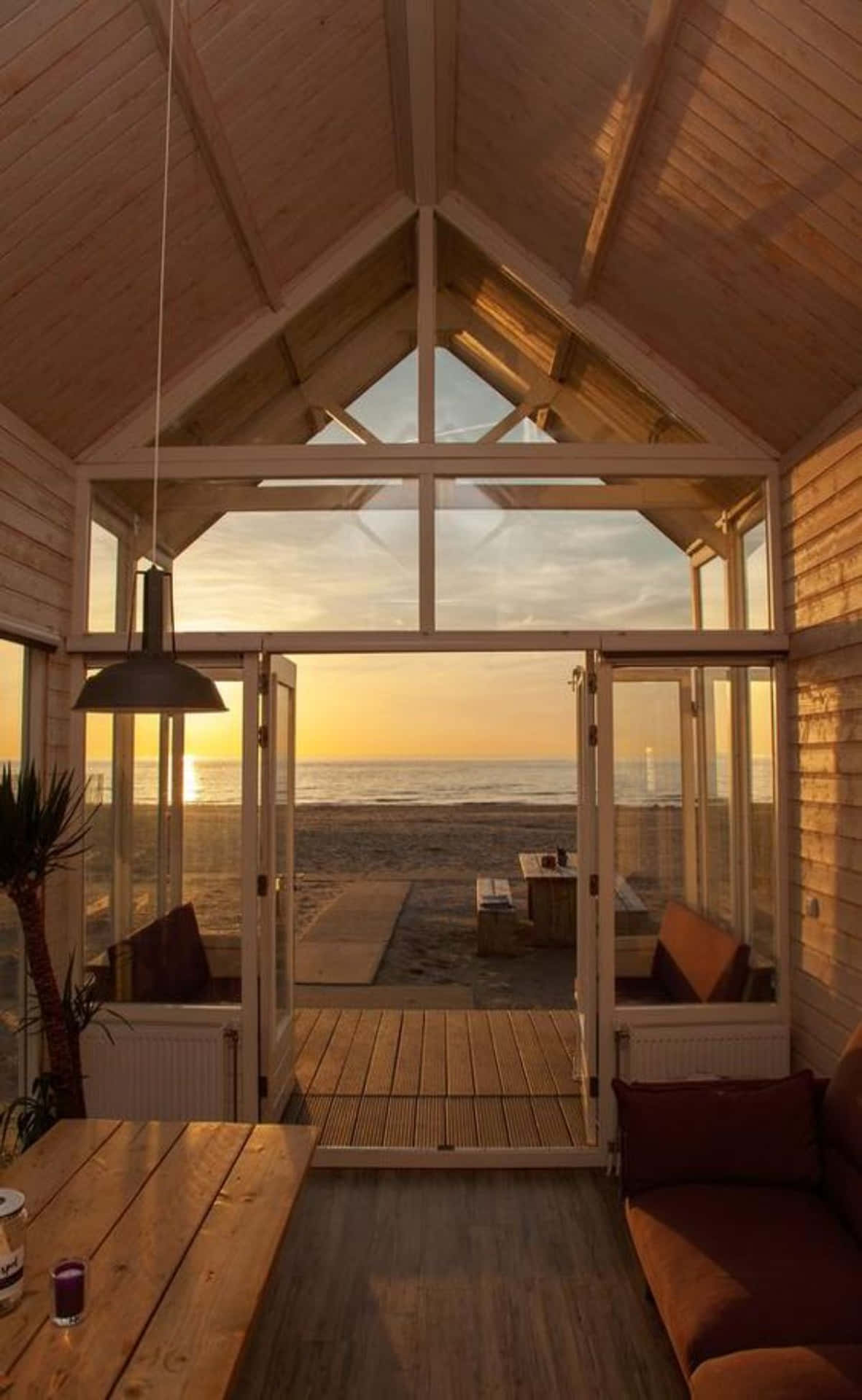 Cottage 桌布 壁紙 — 溫暖的木質小屋內部通向寧靜的海灘，金色夕陽柔和光線透過玻璃門灑入。