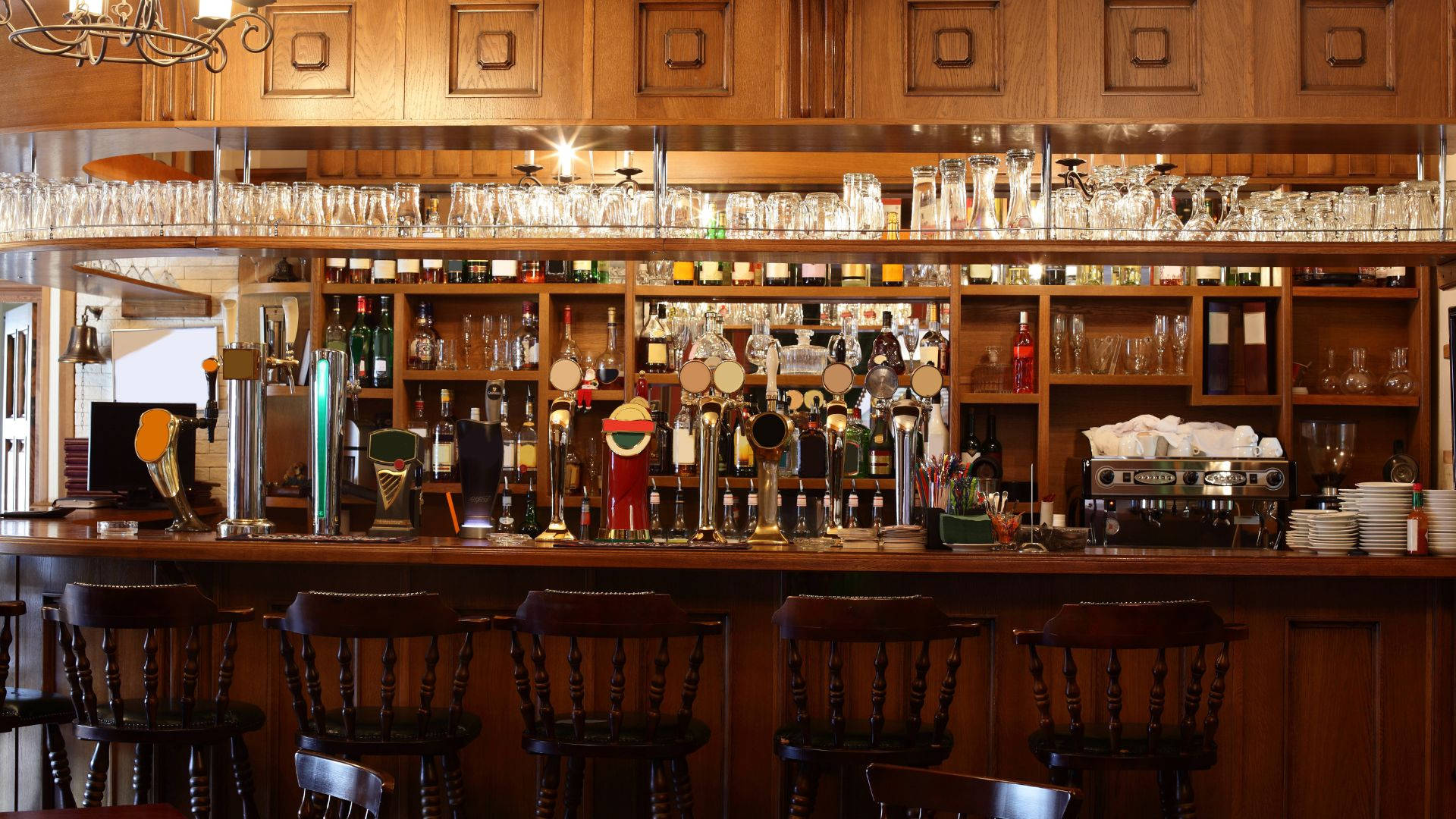 drinks 桌布 壁紙 — 溫暖的木質酒吧，配有拋光凳子、玻璃器皿和啤酒龍頭，在柔和吊燈燈光下。