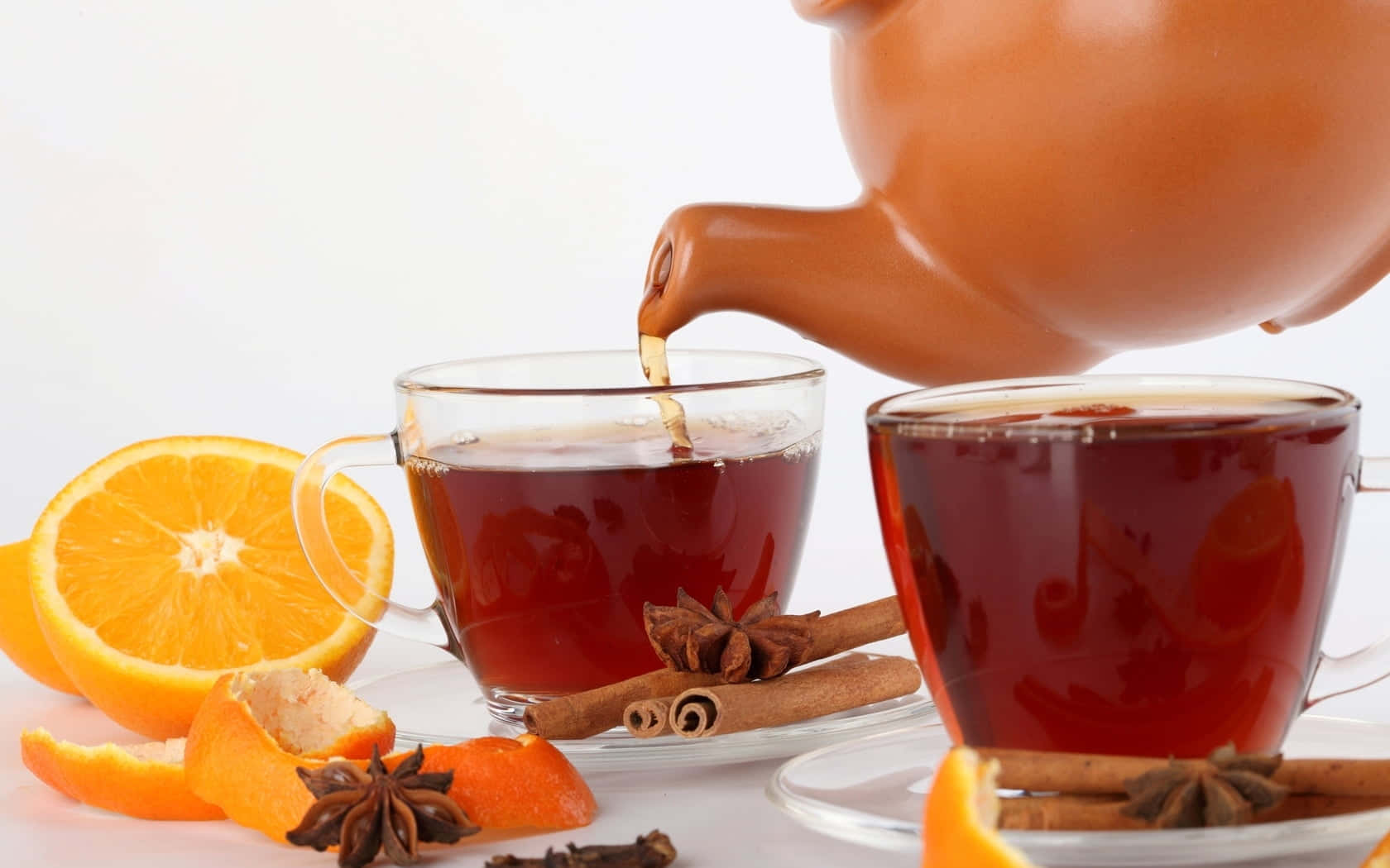 Mulled Wine 桌布 壁紙 — 溫暖的琥珀色茶從陶壺倒入玻璃杯中，周圍有柑橘片、肉桂棒和八角，放在明亮的白色表面上。