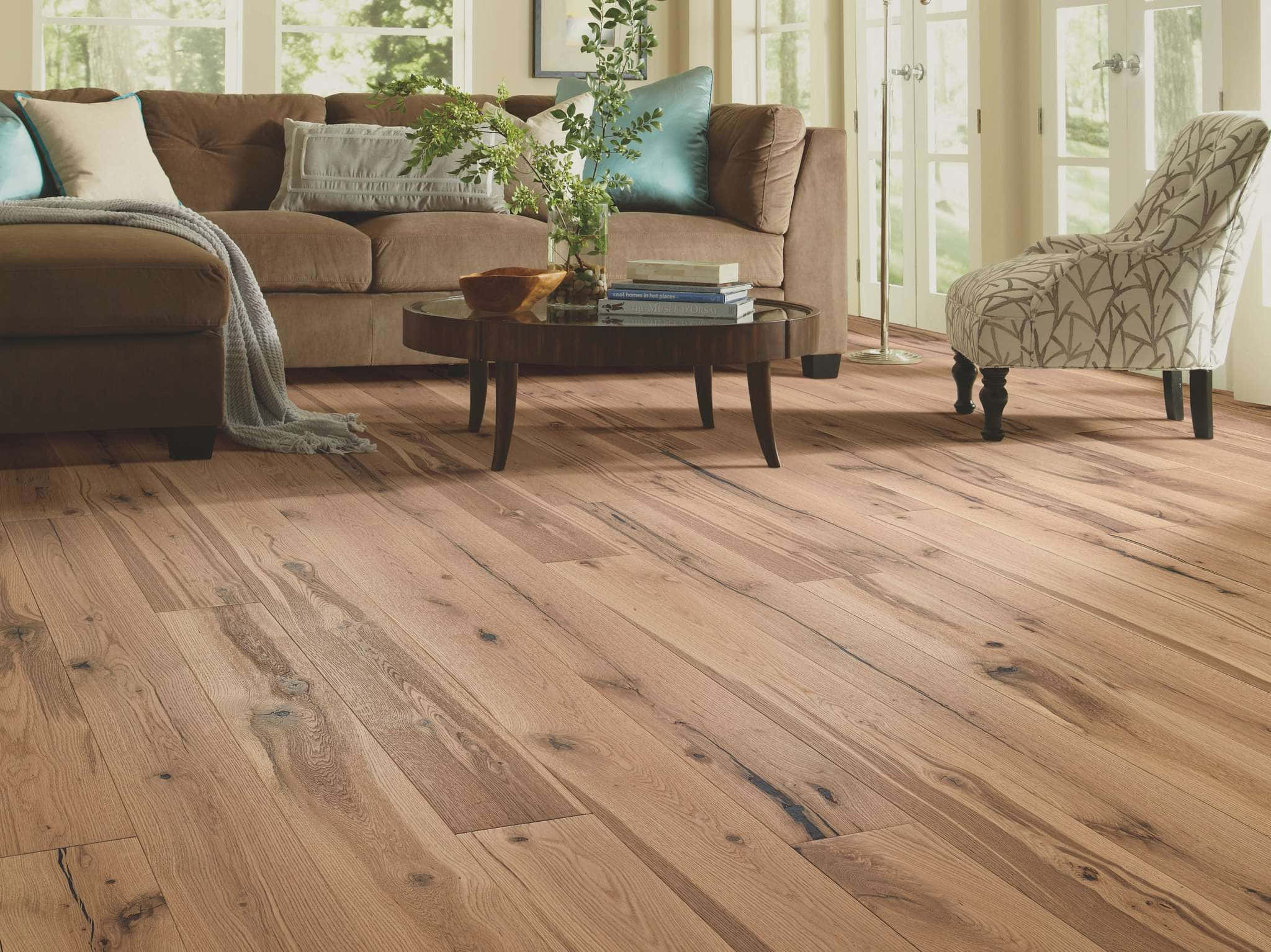 Wood Flooring 桌布 壁紙 — 溫暖的硬木地板延伸穿過一個陽光充足的客廳，配有棕色沙發、青綠色抱枕和圖案扶手椅。