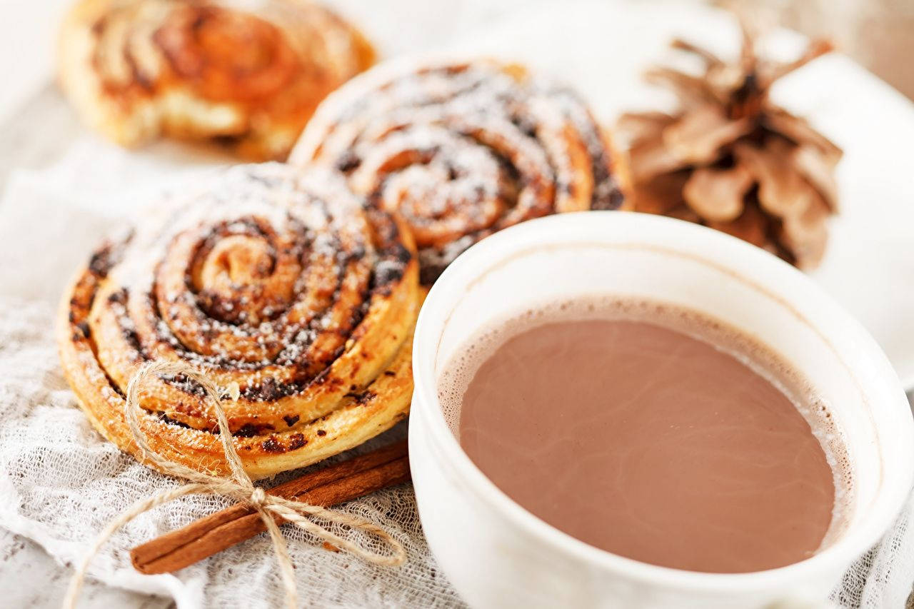 Pastry 桌布 壁紙 — 溫暖的肉桂卷撒上糖，旁邊放著一杯熱巧克力，附有松果和肉桂棒，放在有紋理的布上。