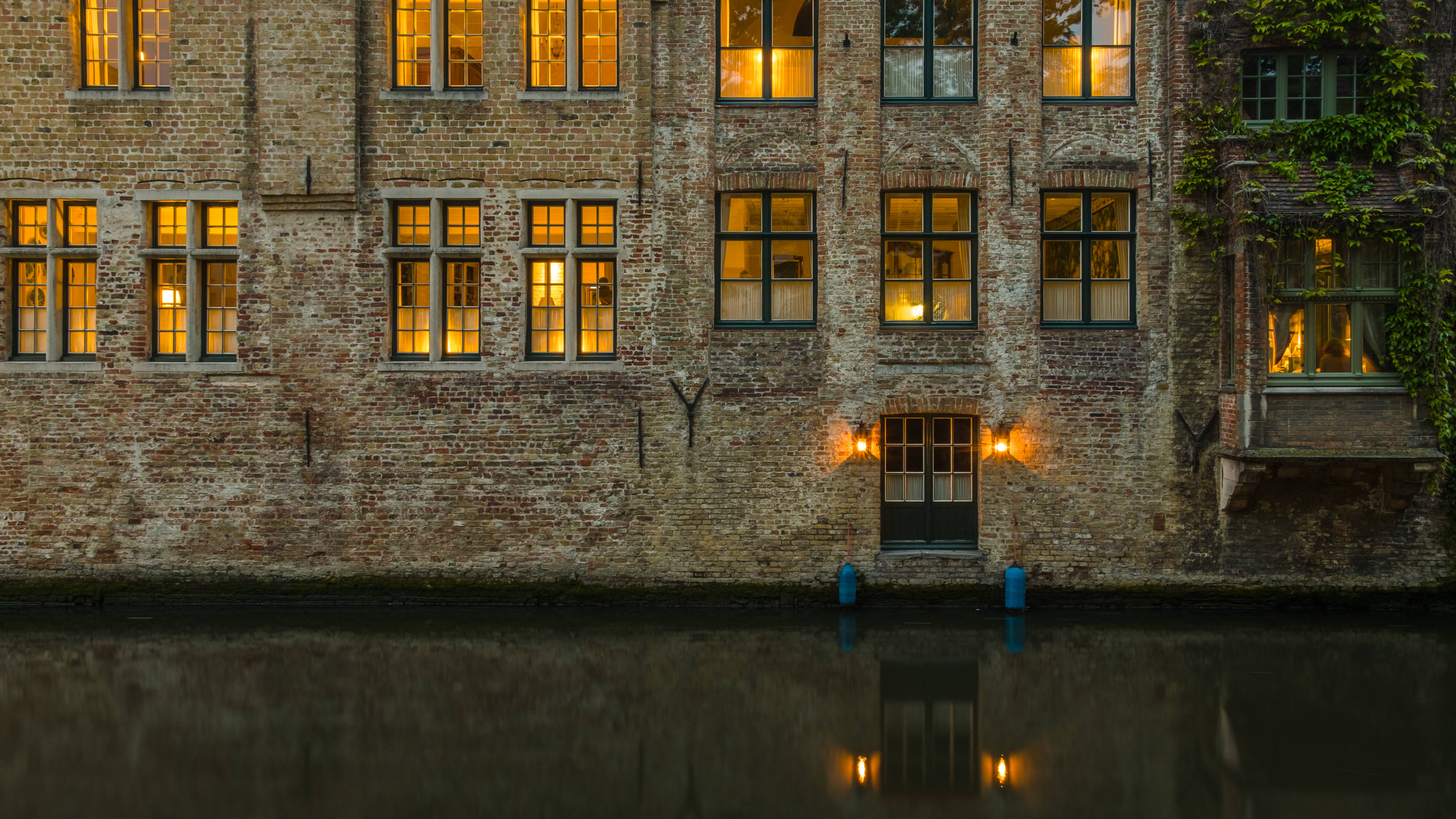 比利時 桌布 壁紙 — 溫暖的金色燈光從一座風化的磚砌建築窗戶中閃耀，倒映在夜晚平靜的運河水面上。