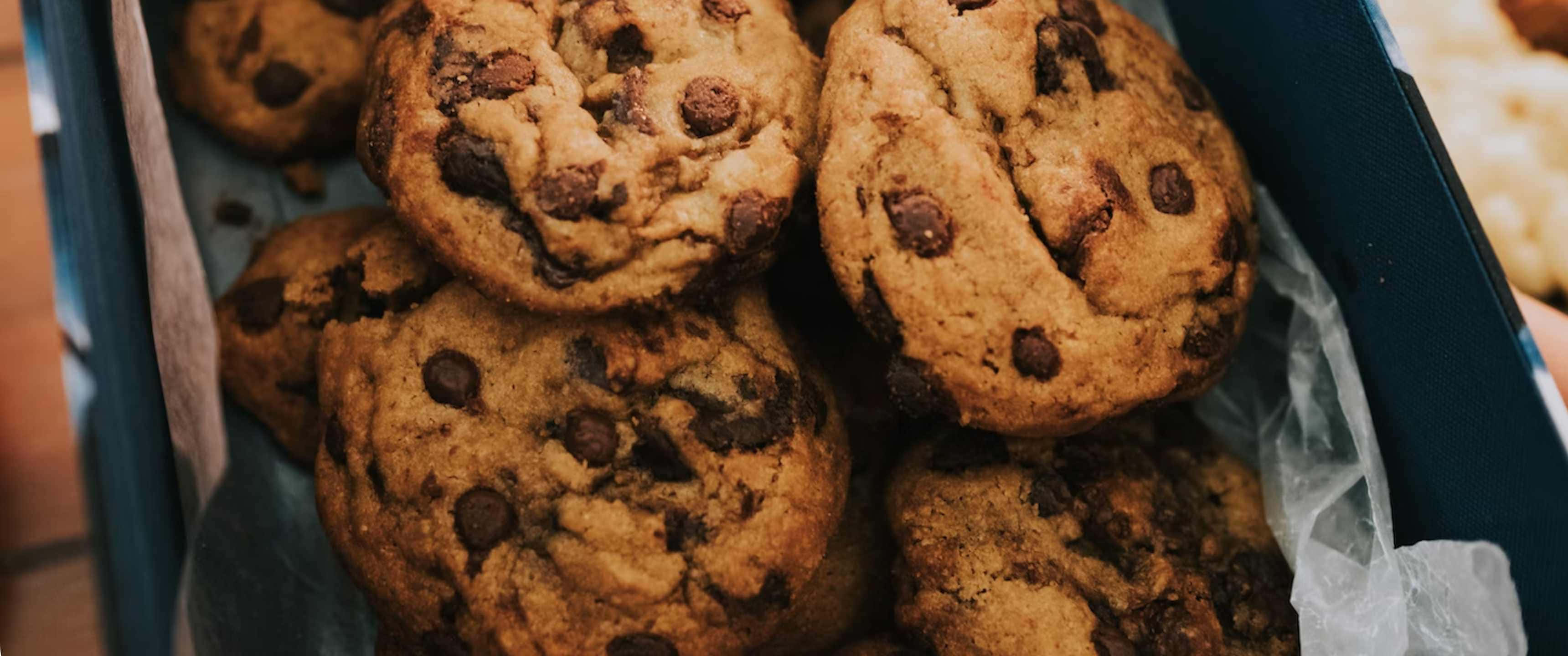 Cookies 桌布 壁紙 — 溫暖的金黃色巧克力餅乾，誘人地堆疊在一個內襯皺紋烘焙紙的深藍色盒子裡。