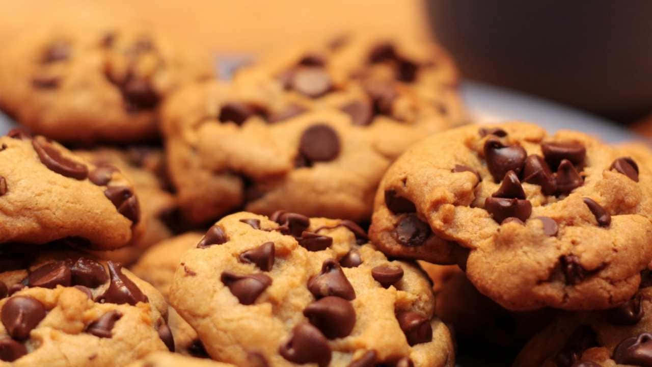 720p Cookies 桌布 壁紙 — 溫暖金黃的巧克力餅乾高高堆起，上面有融化的巧克力豆在柔和光線下閃閃發光。