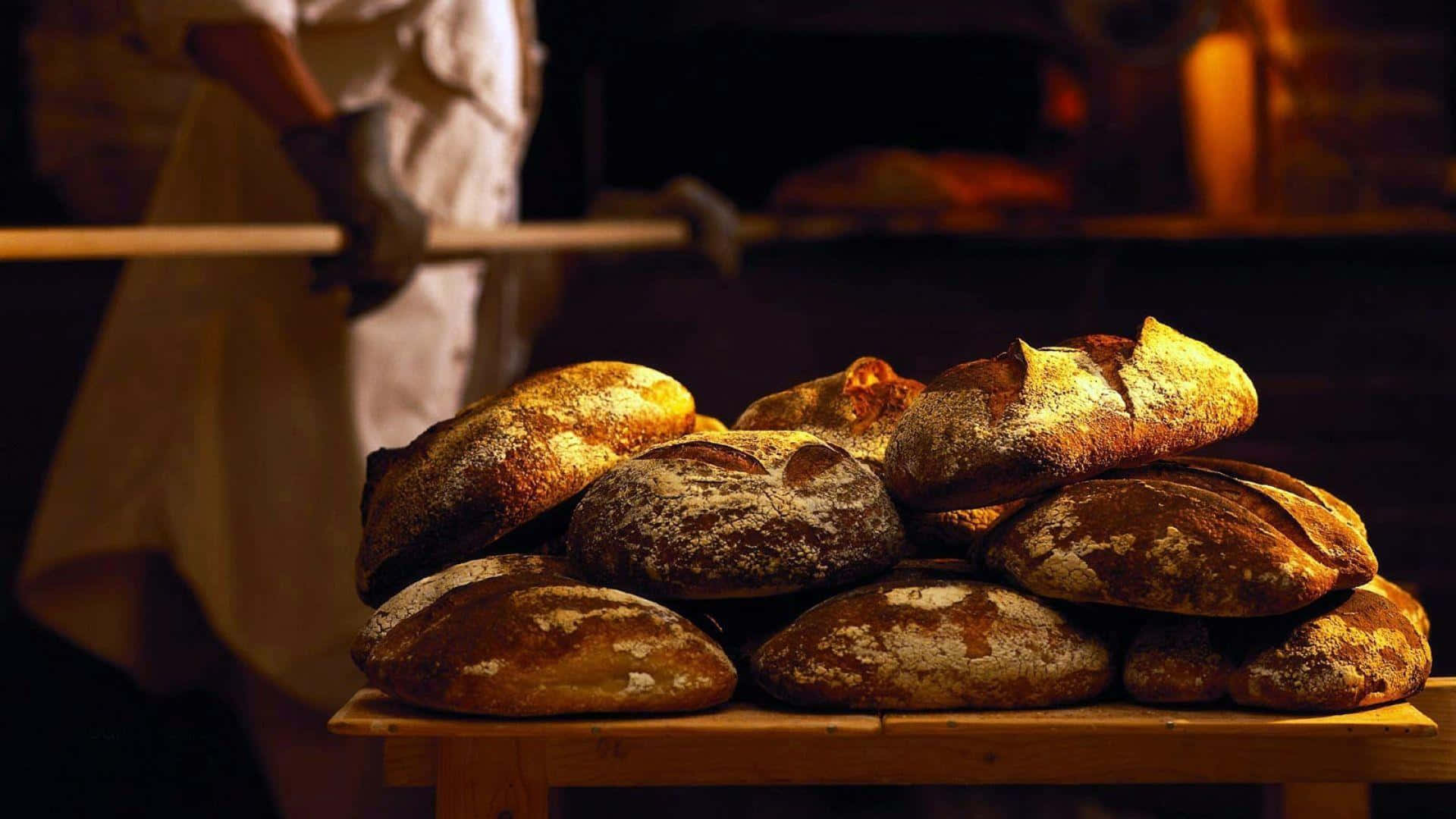 Bakery 桌布 壁紙 — 溫暖金黃色的手工麵包高高堆在木桌上，而麵包師傅正從身後的烤箱中取出新鮮麵包。