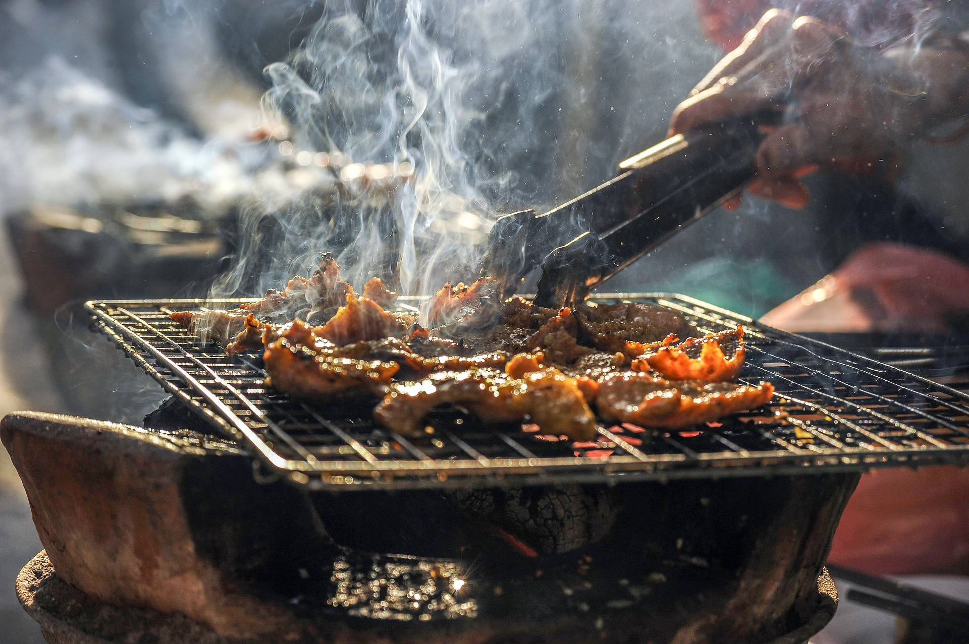 食物動圖 桌布 壁紙 — 滋滋作響的肉在明亮的陽光下冒著濃煙，夾子正在炭火烤架上翻動肉塊。
