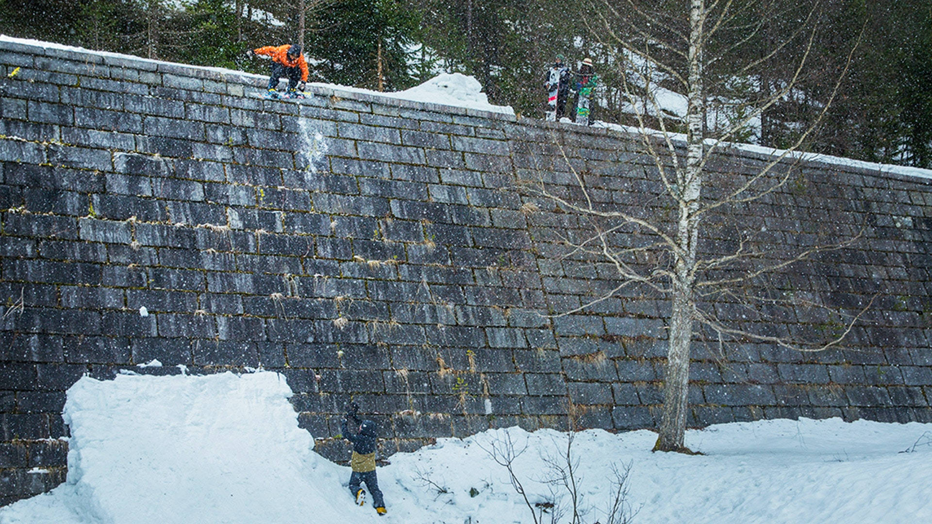 Snowboarding 桌布 壁紙 — 滑雪板運動員在冬季森林中，面對著陡峭的積雪石牆，周圍是光禿禿的樹木和飄落的雪花。