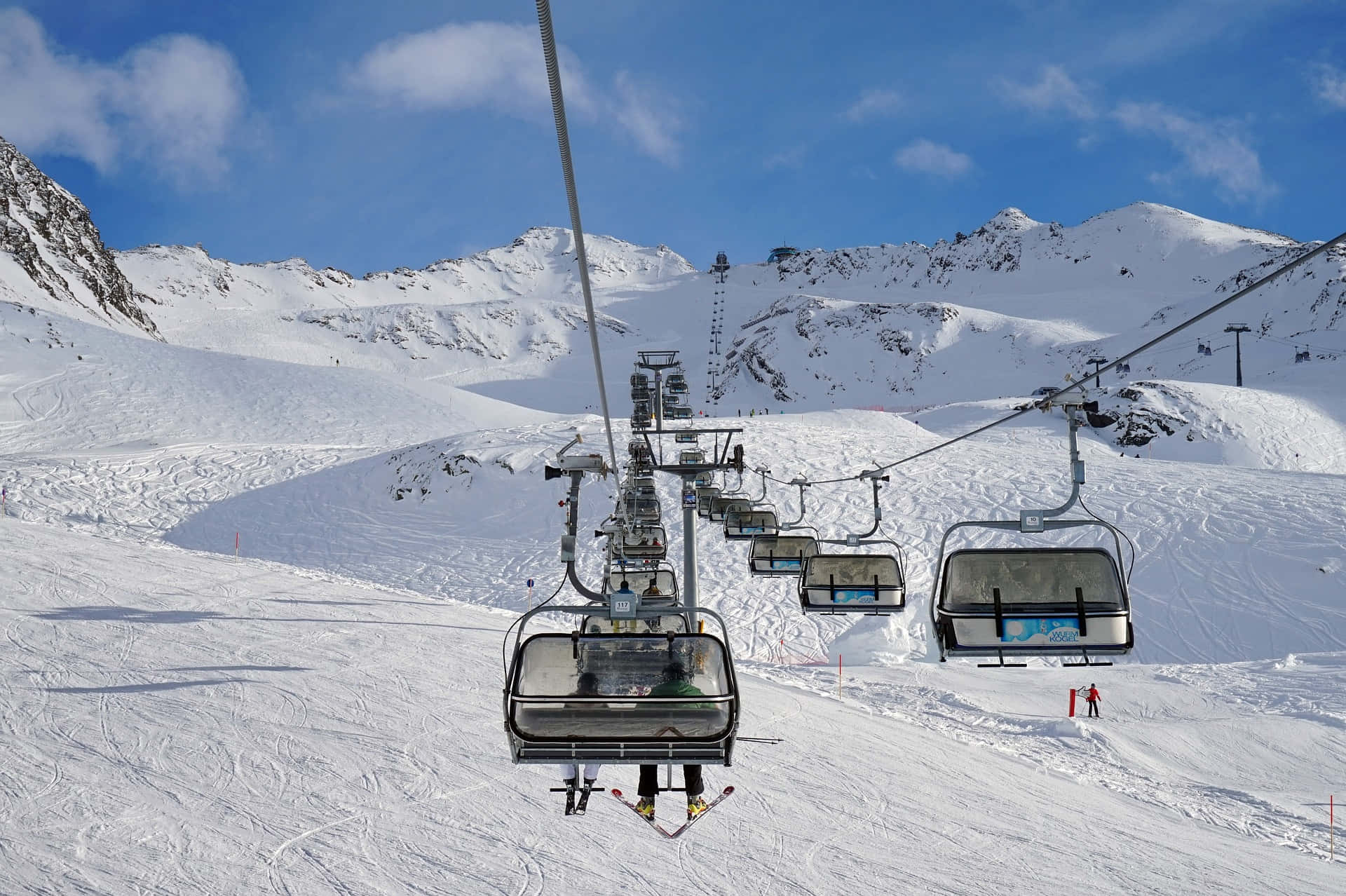 Ski Resort 桌布 壁紙 — 滑雪纜車在晴朗藍天和零星白雲下，攀升於積雪覆蓋的山巒之上。