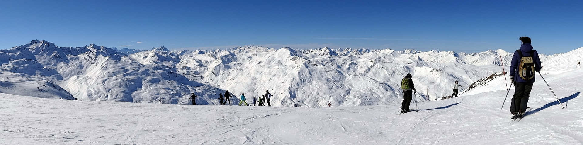 雪 桌布 壁紙 — 滑雪者在一片晴朗藍天下穿越廣闊的雪山山脈，山峰尖銳，陰影深邃。