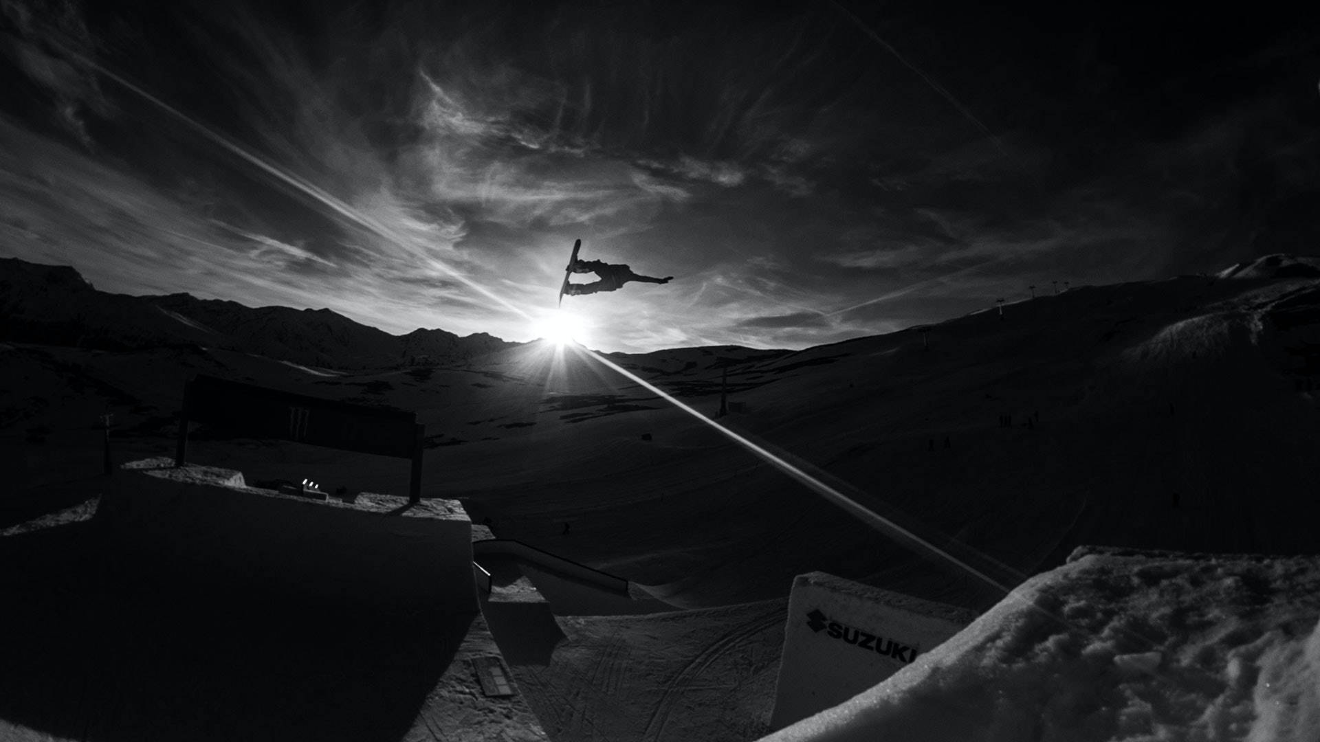 Snowboarding 桌布 壁紙 — 滑雪運動員在白雪覆蓋的山峰上，以戲劇性的日出為背景，在空中翱翔，呈現高對比度的黑白畫面。