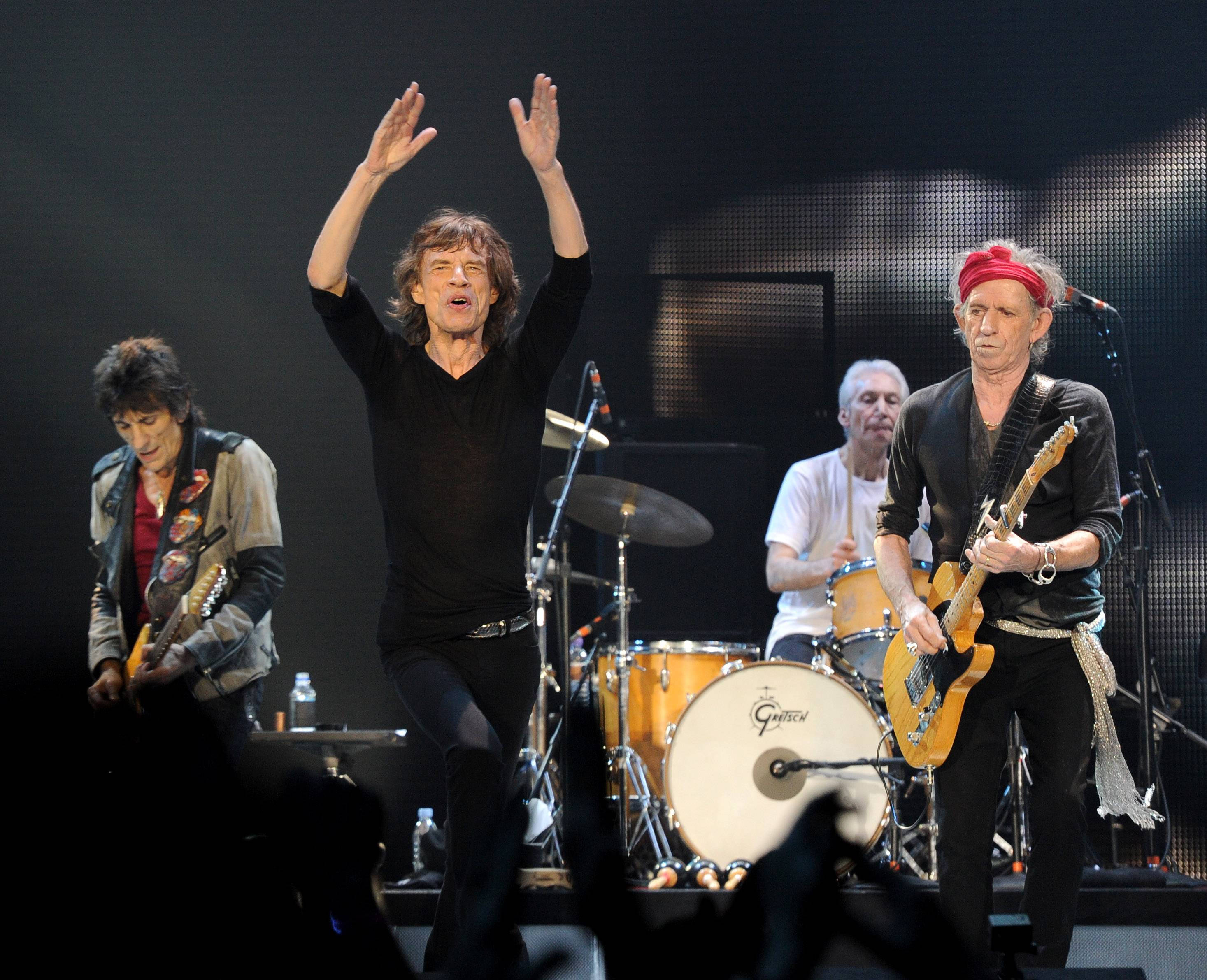 Rock 桌布 壁紙 — 滾石樂團在舞台上充滿活力地表演，米克·賈格爾舉起雙臂，基思·理查茲彈奏吉他，鼓手在Gretsch鼓組後面，在明亮的舞台燈光下。
