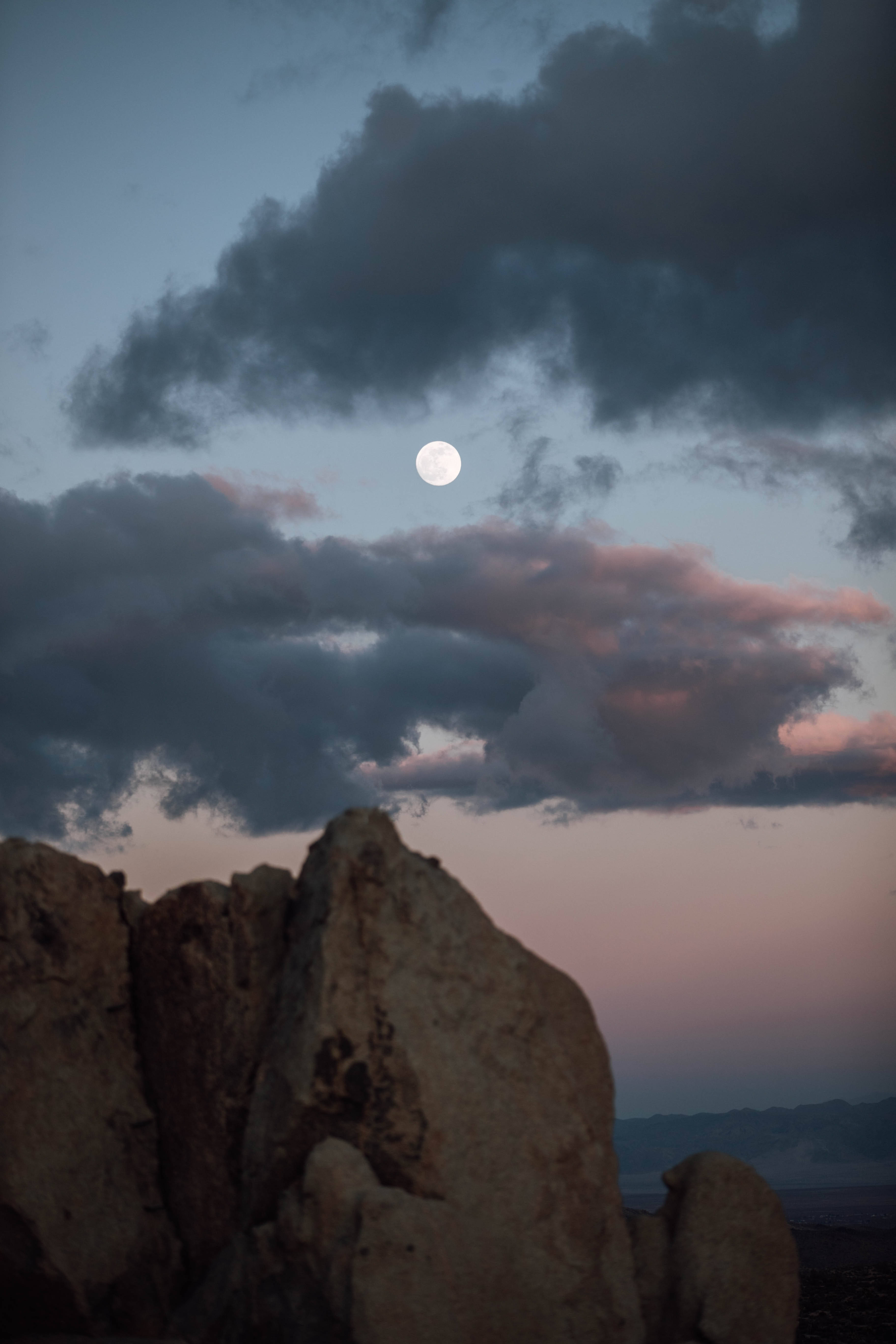 full moon 桌布 壁紙 — 滿月在暮色中透過戲劇性的雲層照耀在崎嶇的沙漠岩石 formations 上。