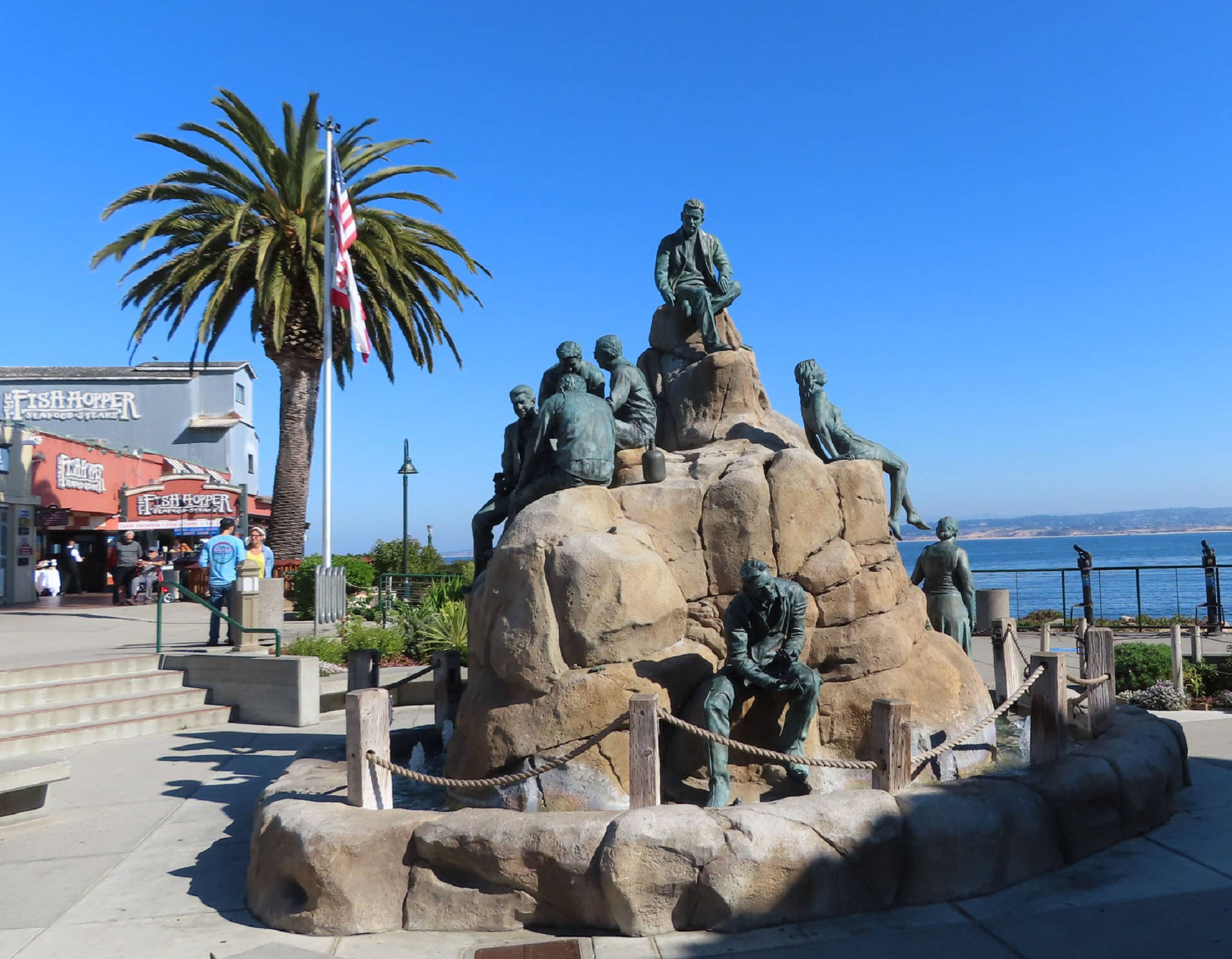 Statue Aesthetic 桌布 壁紙 — 漁夫的銅像棲息在岩石噴泉上，在晴朗的藍天下，旁邊有棕櫚樹和海景。