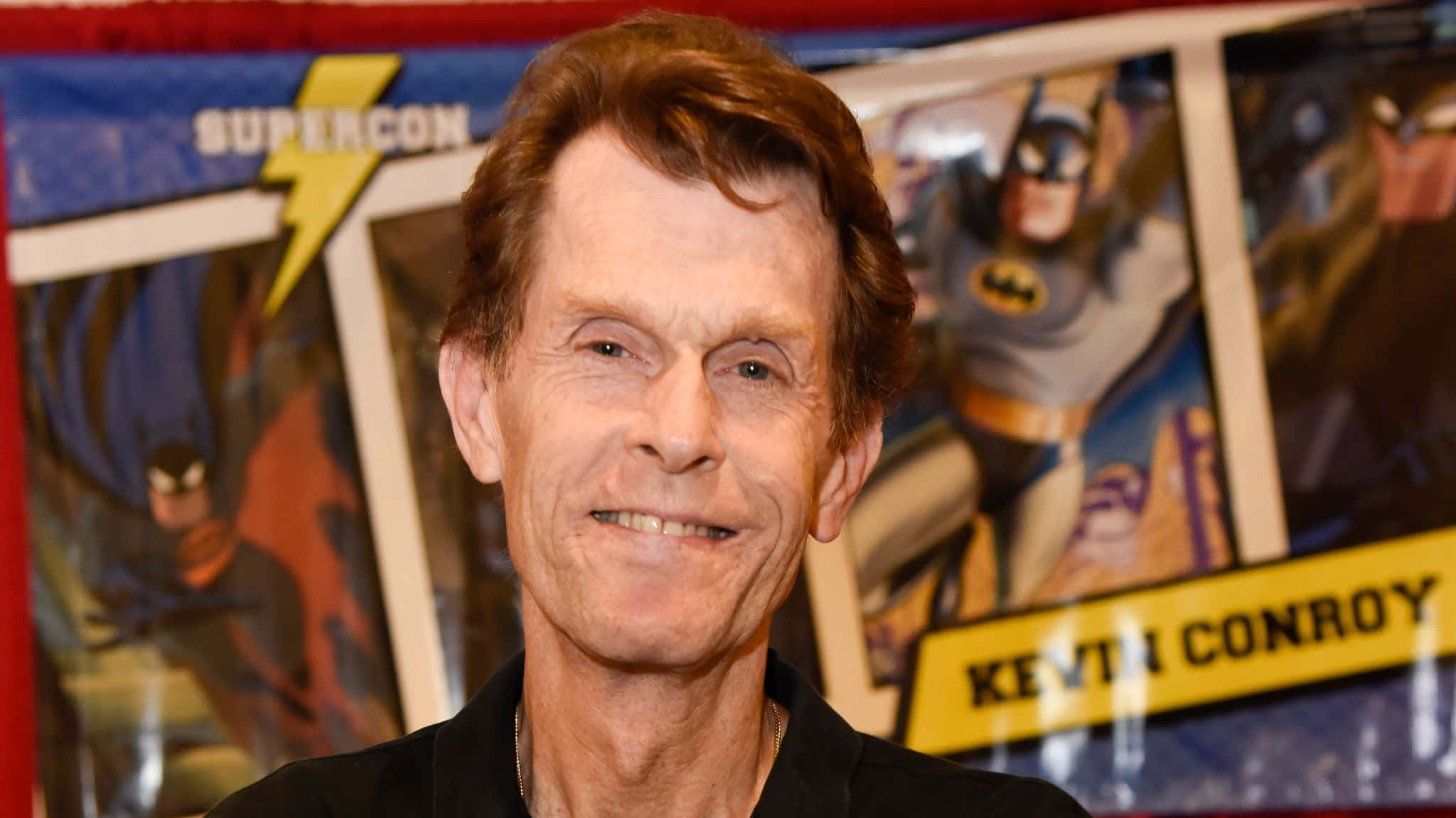 Kevin Conroy 桌布 壁紙 — 演員 Kevin Conroy 在 SuperCon 活動中溫暖微笑，身後有蝙蝠俠海報。